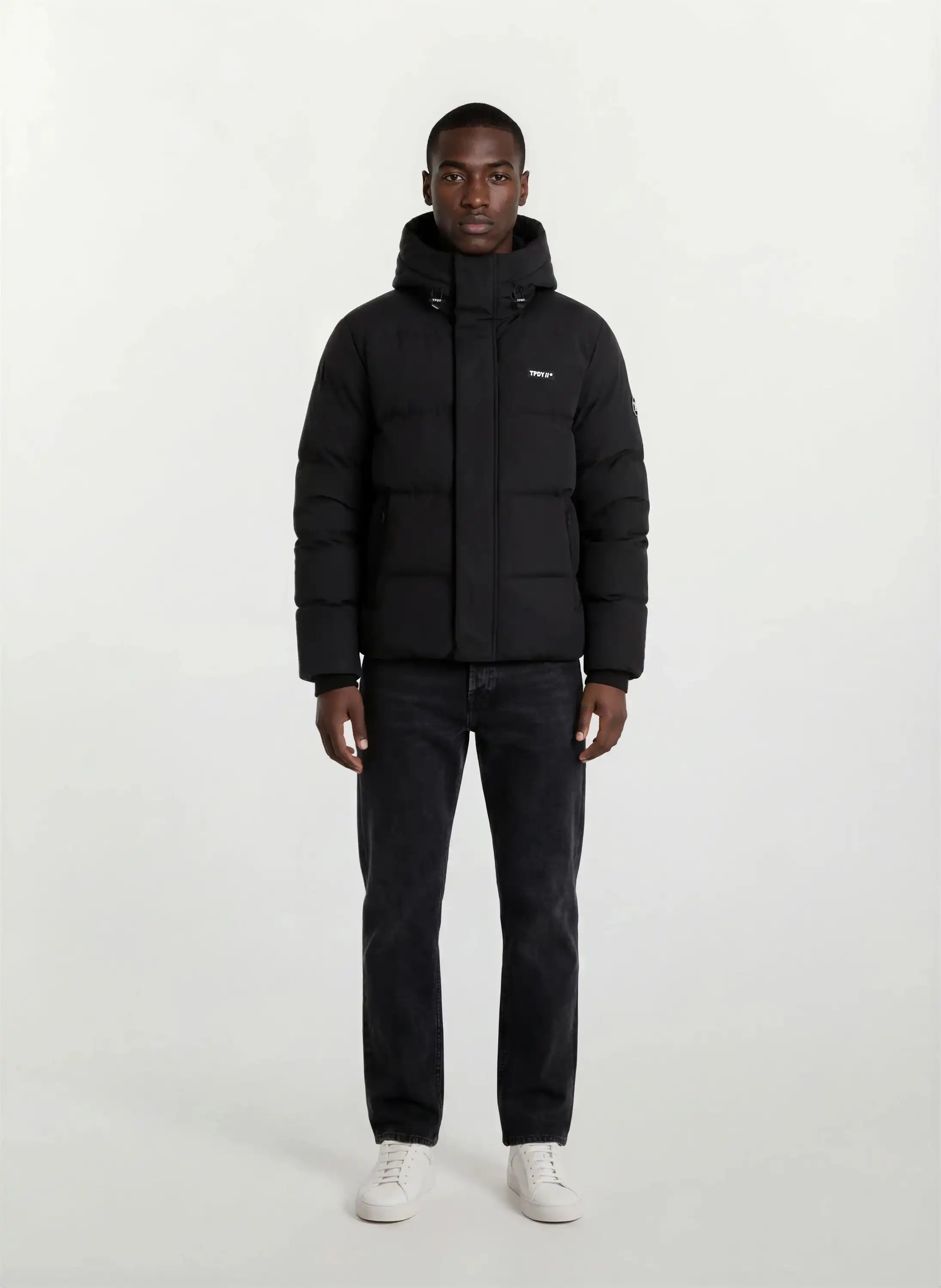 Eine Person steht mit dem Gesicht nach vorne vor einem schlichten weißen Hintergrund und trägt die trueprodigy Herren Winterjacke mit fester Kapuze im Oversized Fit, gepaart mit dunklen Jeans und weißen Sneakers.