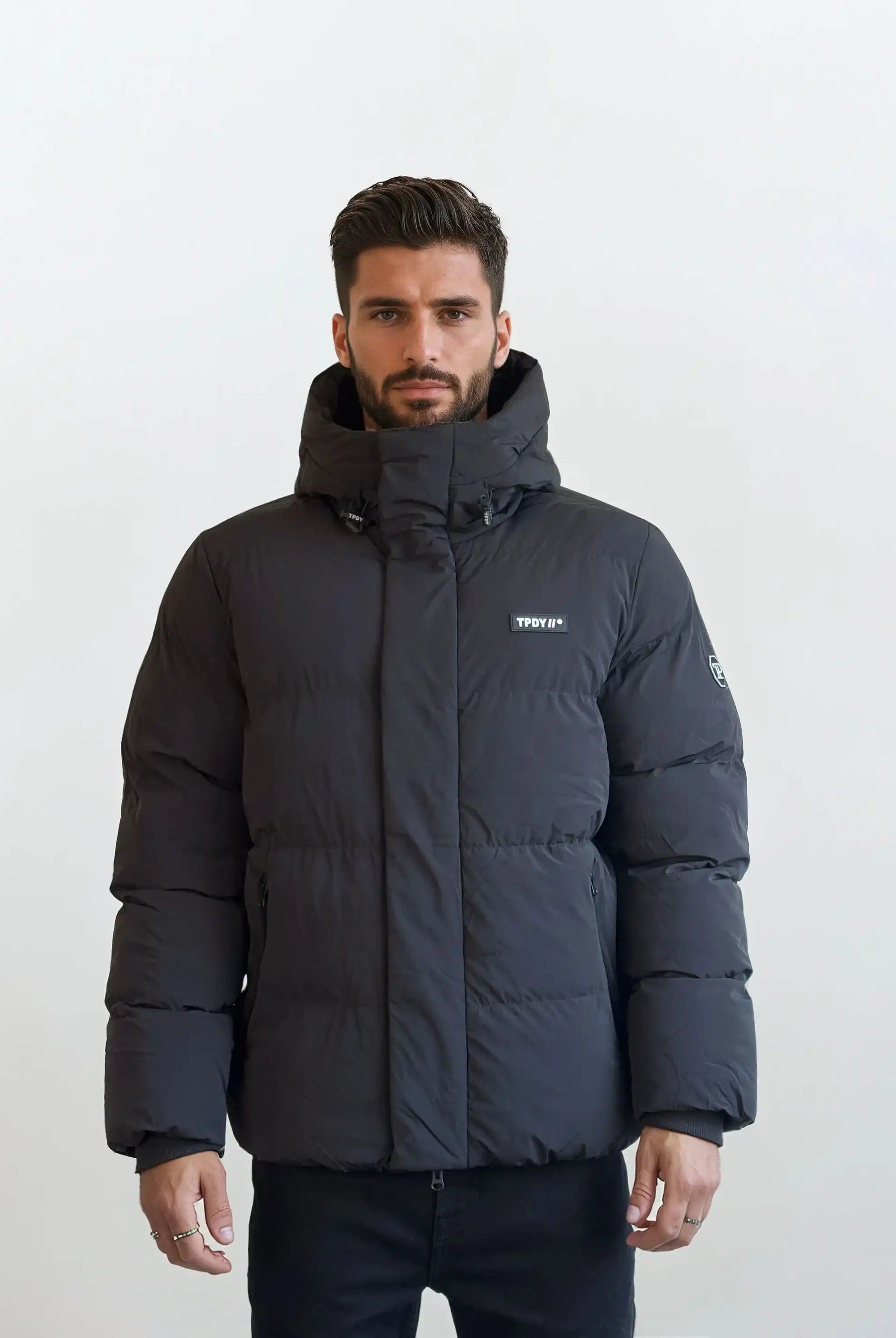 Ein Mann mit kurzem dunklem Haar und Bart trägt eine schwarze trueprodigy Herren Winterjacke mit fester Kapuze im Oversized Fit, wasserabweisend, mit kleinem Logo auf der Brust und Patch am linken Ärmel. Weißer Hintergrund.