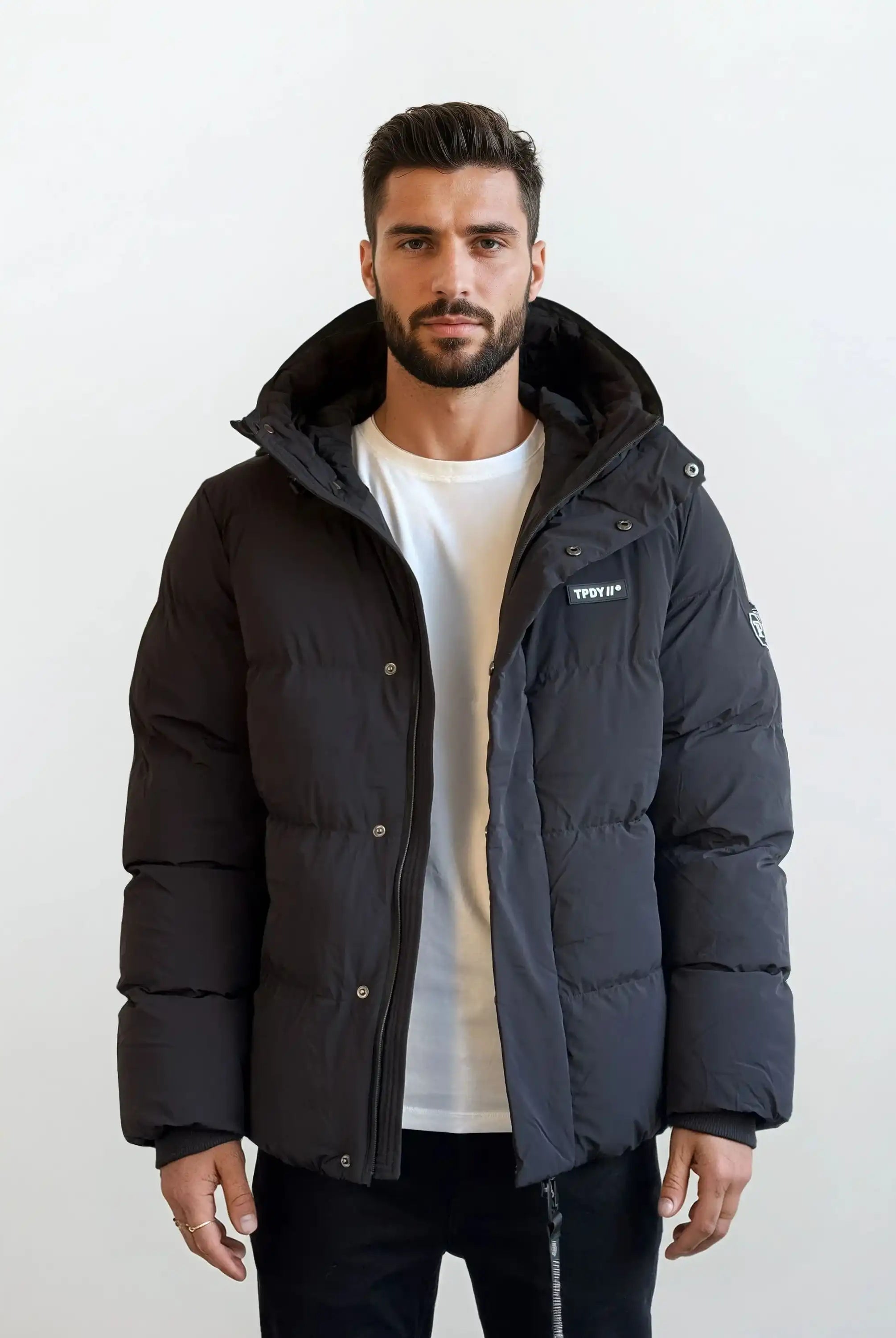 Ein Mann mit kurzen dunklen Haaren und einem Bart steht vor einem schlichten weißen Hintergrund und trägt eine trueprodigy Herren Winterjacke mit fester Kapuze im Oversized Fit über einem weißen T-Shirt und schwarzen Hosen. Die Jacke trägt einen kleinen Patch auf der linken Brust.