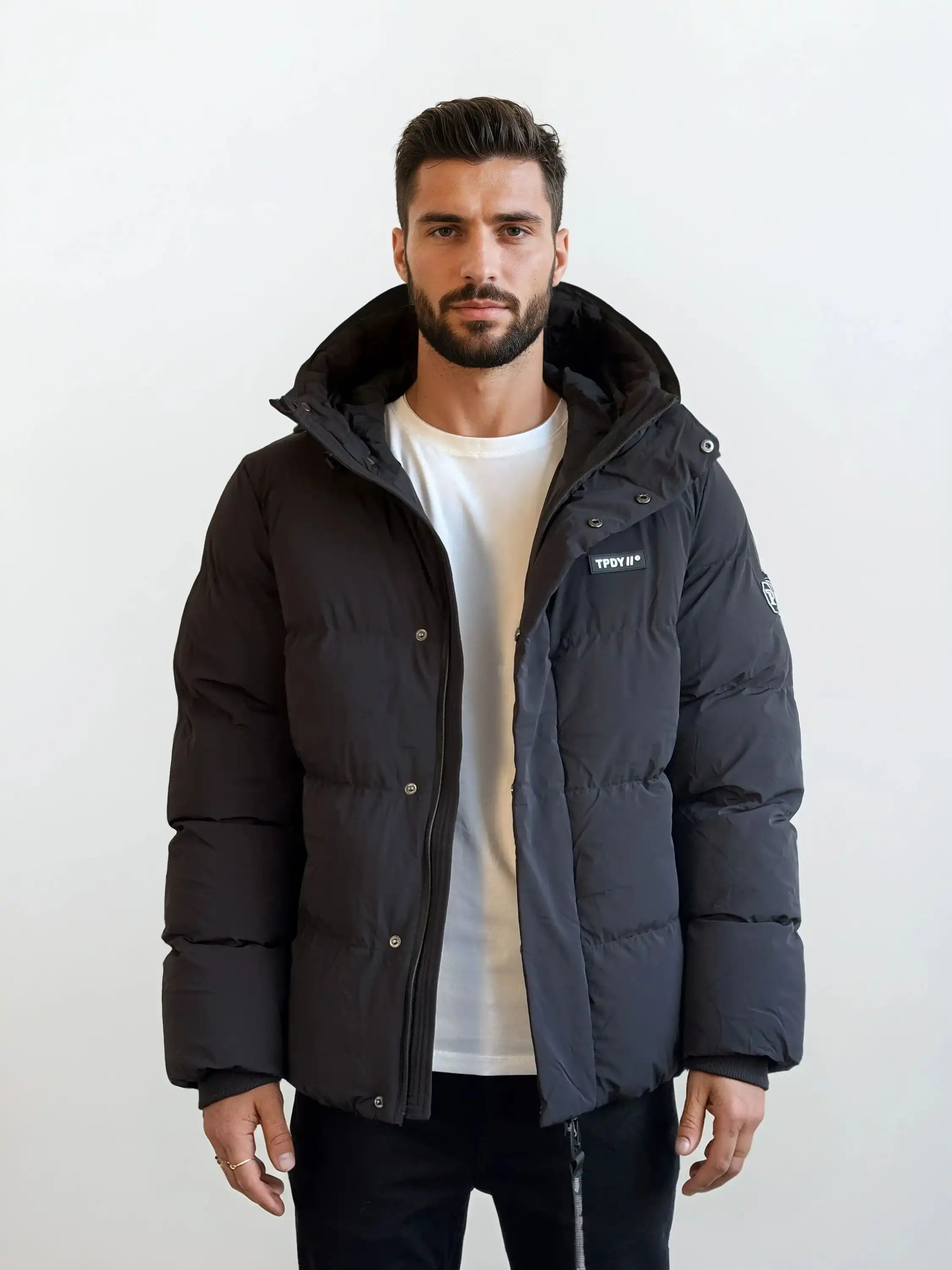 Ein Mann mit kurzen dunklen Haaren und einem Bart steht vor einem schlichten weißen Hintergrund und trägt eine trueprodigy Herren Winterjacke mit fester Kapuze im Oversized Fit über einem weißen T-Shirt und schwarzen Hosen. Die Jacke trägt einen kleinen Patch auf der linken Brust.