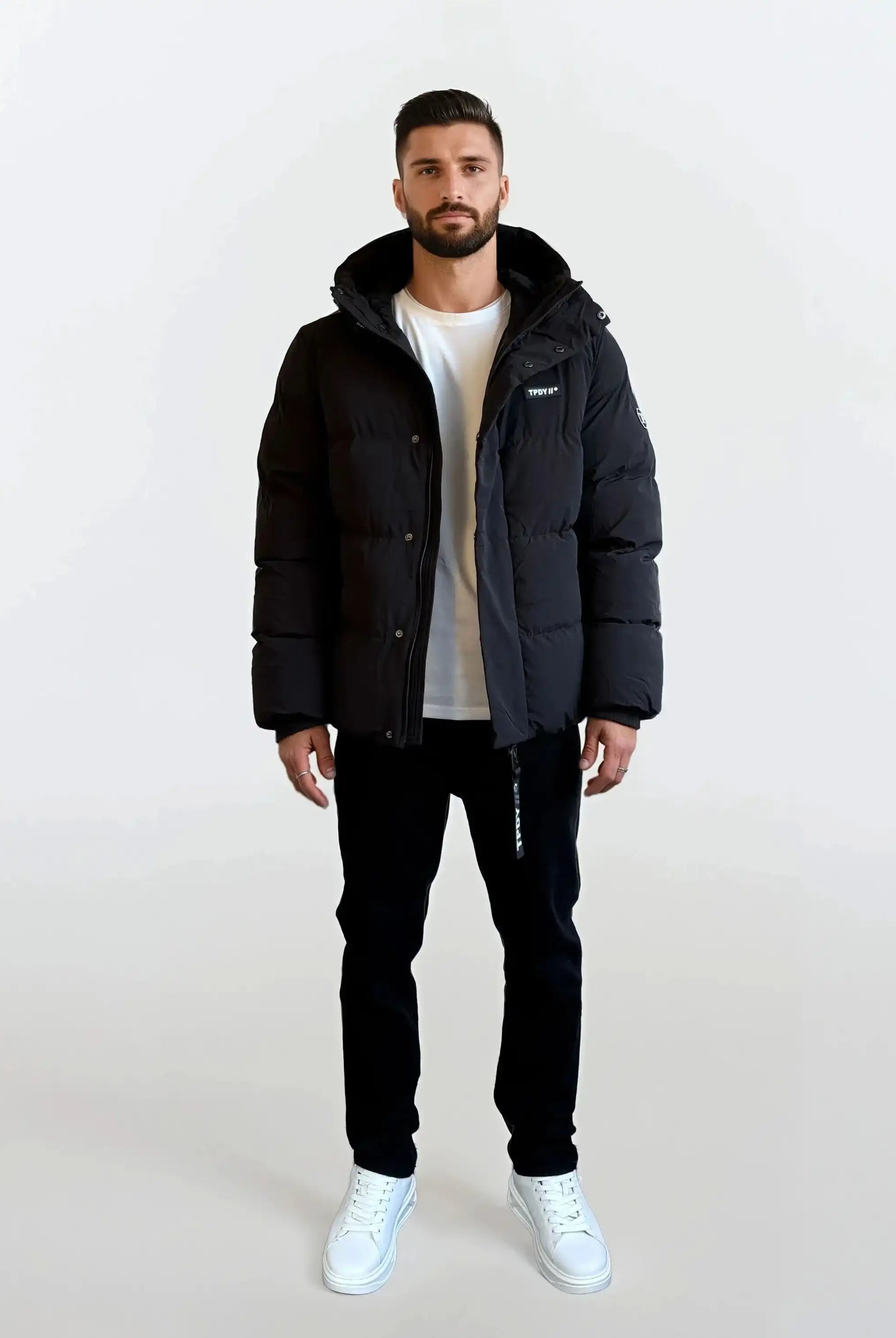 Ein Mann steht mit dem Gesicht nach vorne vor einem schlichten weißen Hintergrund und trägt die trueprodigy Herren Winterjacke mit fester Kapuze im Oversized Fit, ein weißes T-Shirt, eine schwarze Hose und weiße Turnschuhe.