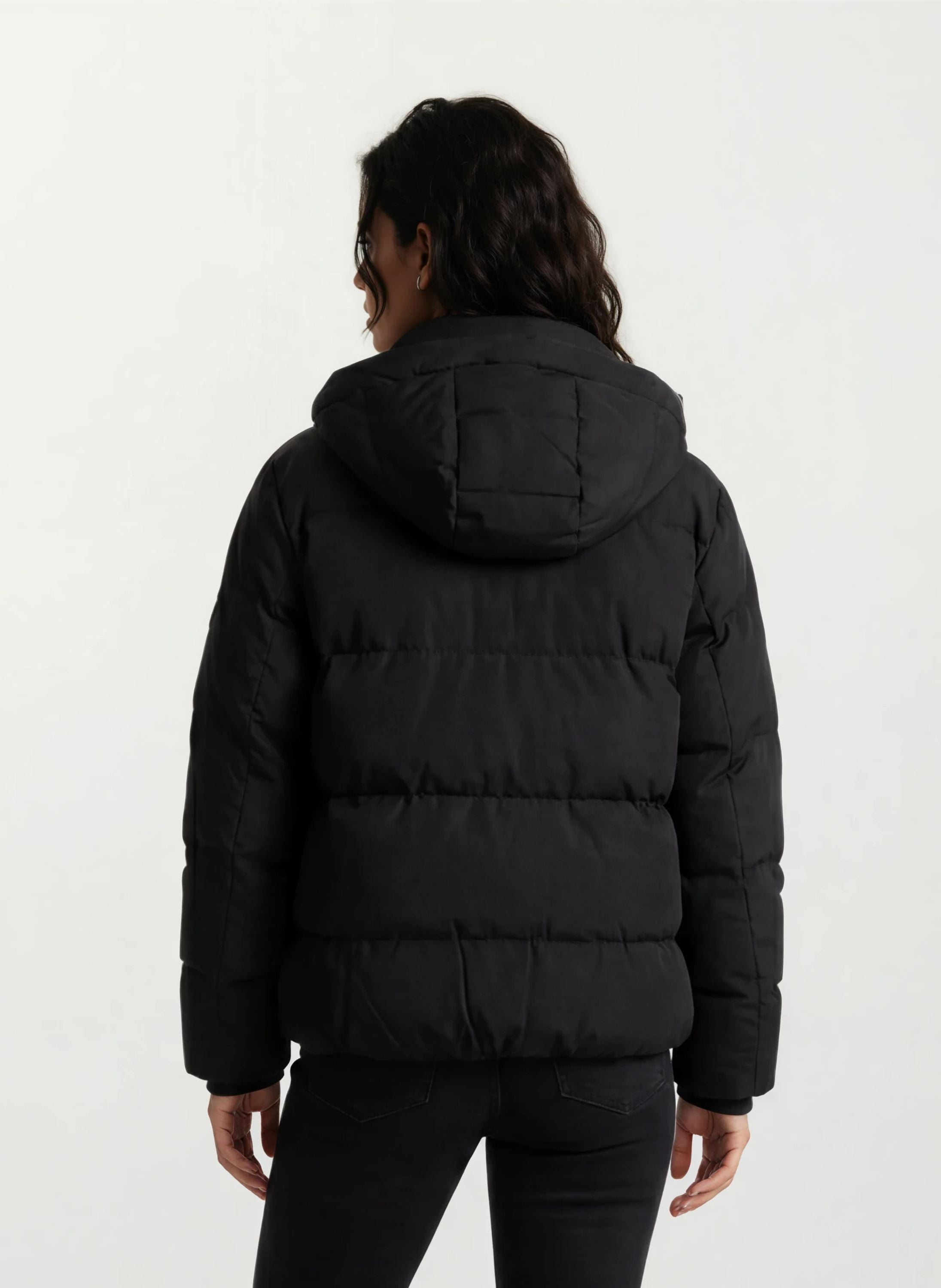 Eine Person steht mit dem Rücken zur Kamera und trägt die trueprodigy Damen Winterjacke mit fester Kapuze im Oversized Fit und schwarzer Hose vor einem einfachen hellen Hintergrund.