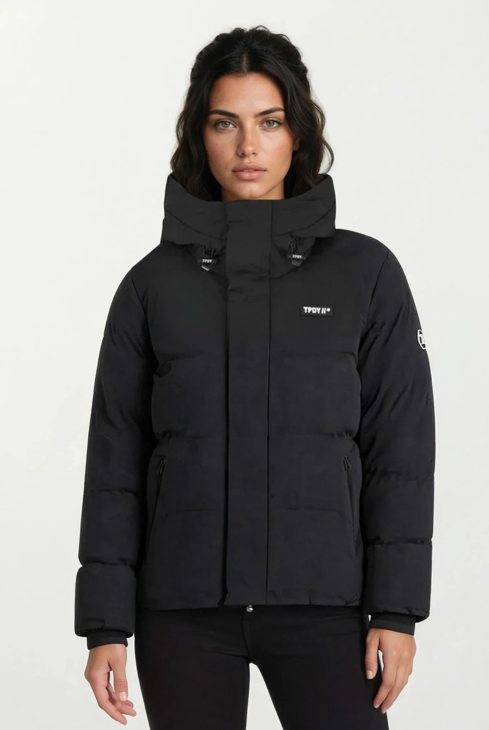 Eine Frau mit langen dunklen Haaren trägt eine trueprodigy Damen Winterjacke im Oversized Fit mit fester Kapuze und hoher wasserabweisender Funktion, kleinen weißen Logos auf Brust und linkem Ärmel vor hellem Hintergrund.
