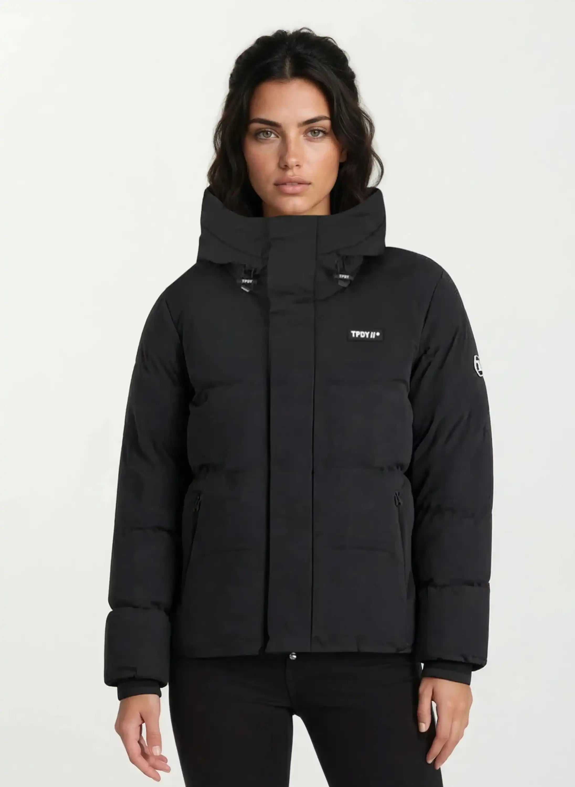 Eine Frau mit langen dunklen Haaren trägt eine trueprodigy Damen Winterjacke im Oversized Fit mit fester Kapuze und hoher wasserabweisender Funktion, kleinen weißen Logos auf Brust und linkem Ärmel vor hellem Hintergrund.