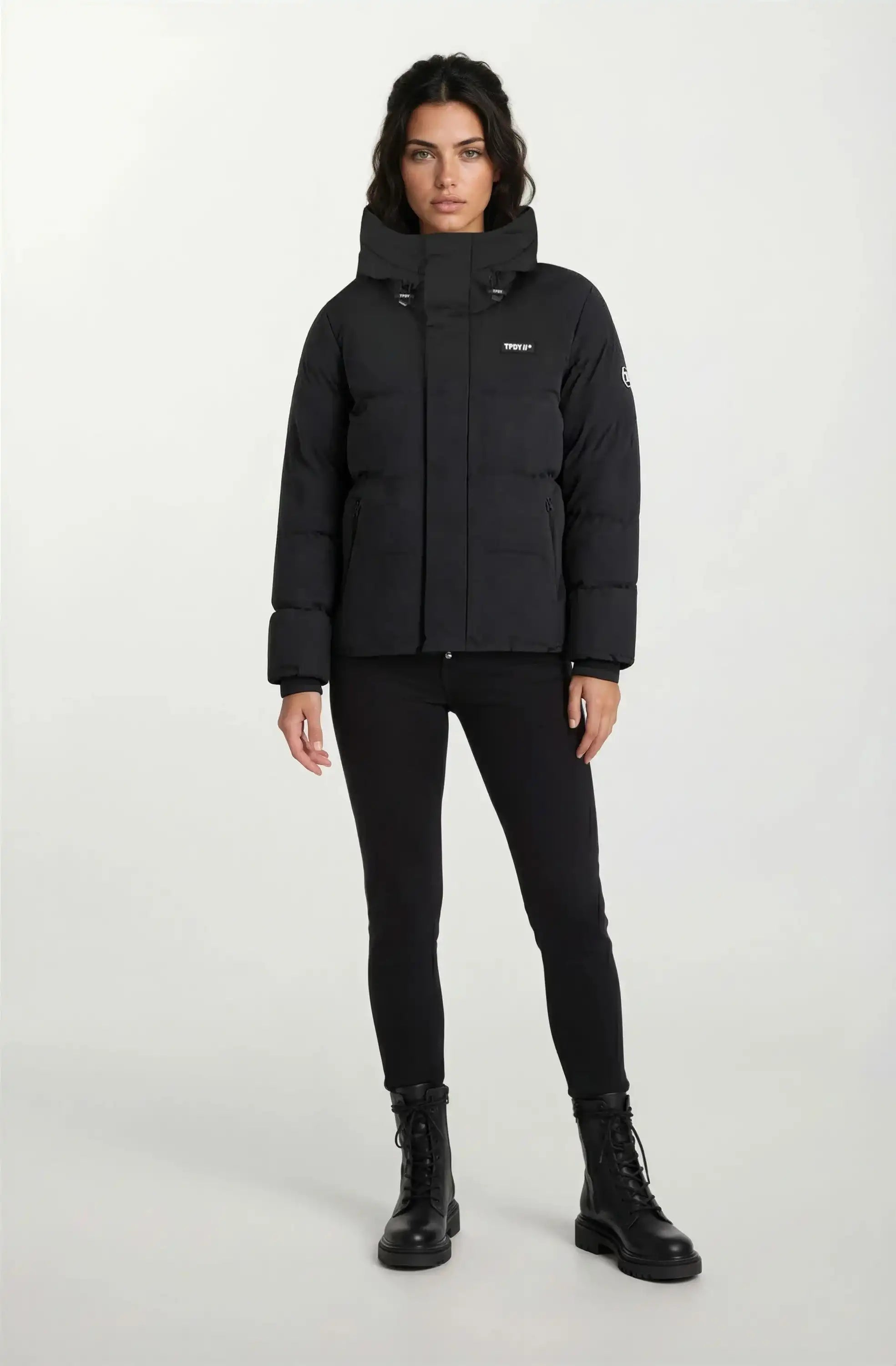 Eine Person steht frontal vor weißem Hintergrund und trägt eine trueprodigy Damen Winterjacke mit fester Kapuze im Oversized Fit, schwarze Skinny Pants und schwarze Lace-up Boots.