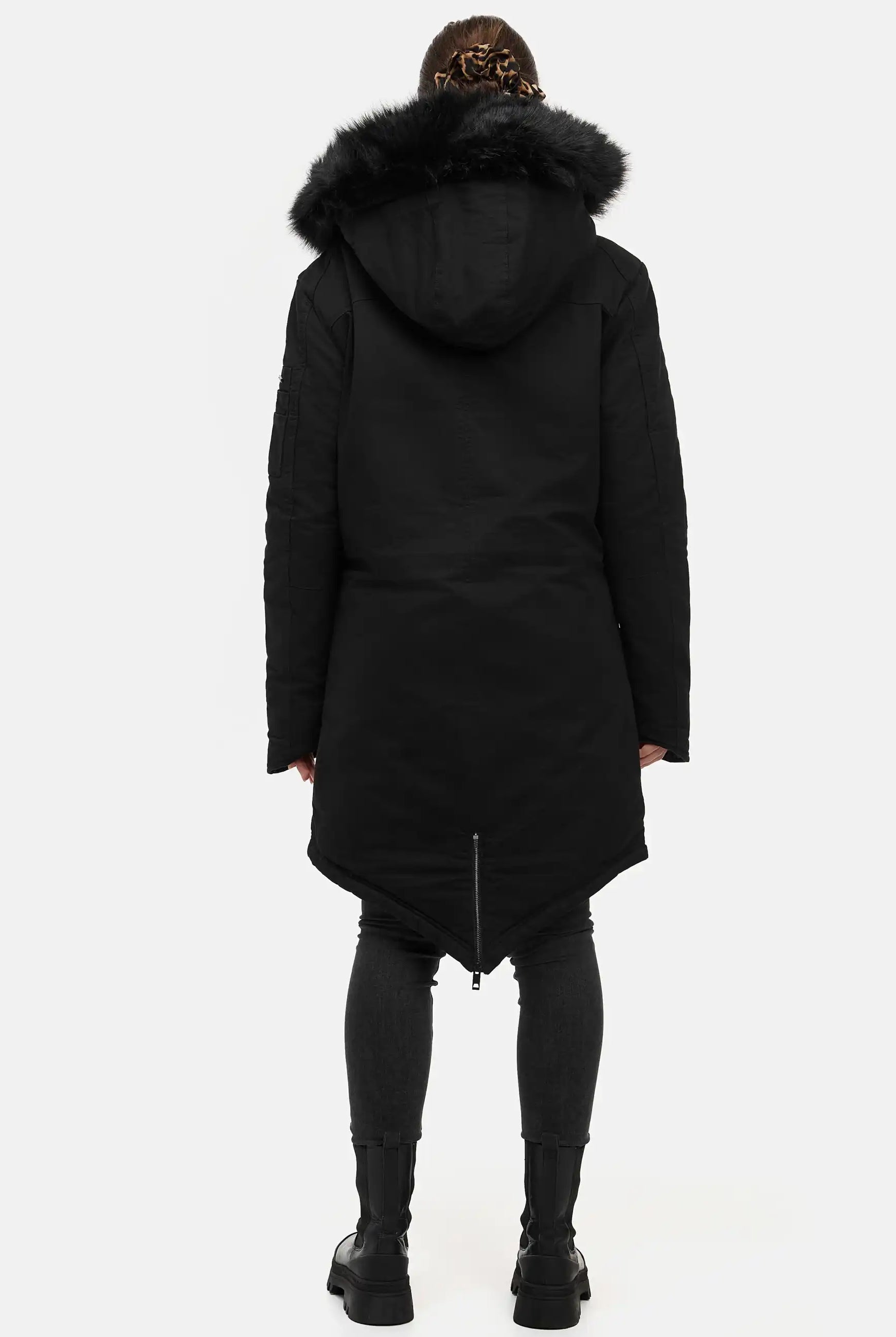 Eine Person blickt von der Kamera weg und trägt den trueprodigy Harvey F Damenparka mit Kapuze und Kunstfell, eine taillierte dunkle Hose und schwarze Stiefel. Der Hintergrund ist einfarbig weiß.