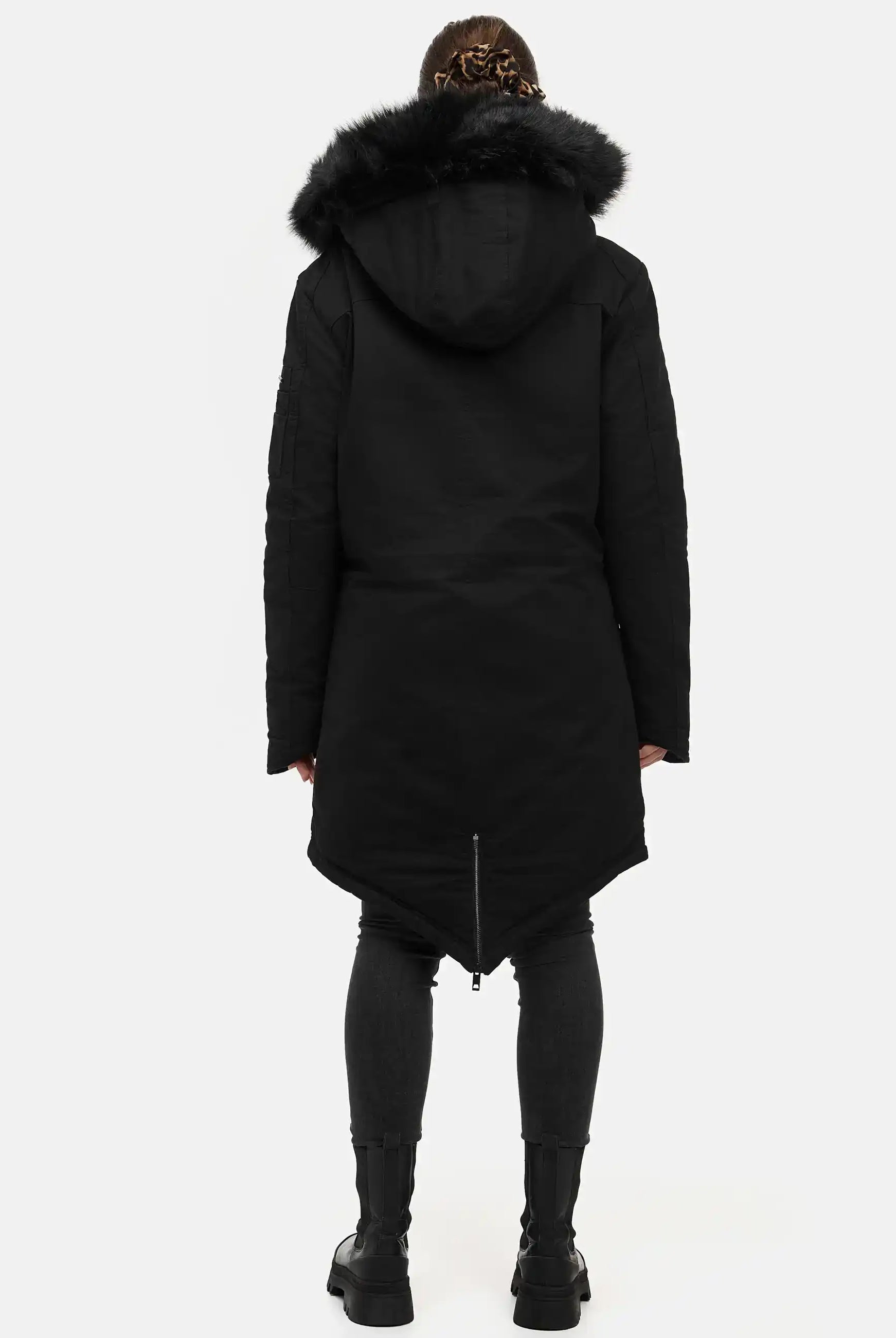 Eine Person blickt von der Kamera weg und trägt den trueprodigy Harvey F Damenparka mit Kapuze und Kunstfell, eine taillierte dunkle Hose und schwarze Stiefel. Der Hintergrund ist einfarbig weiß.