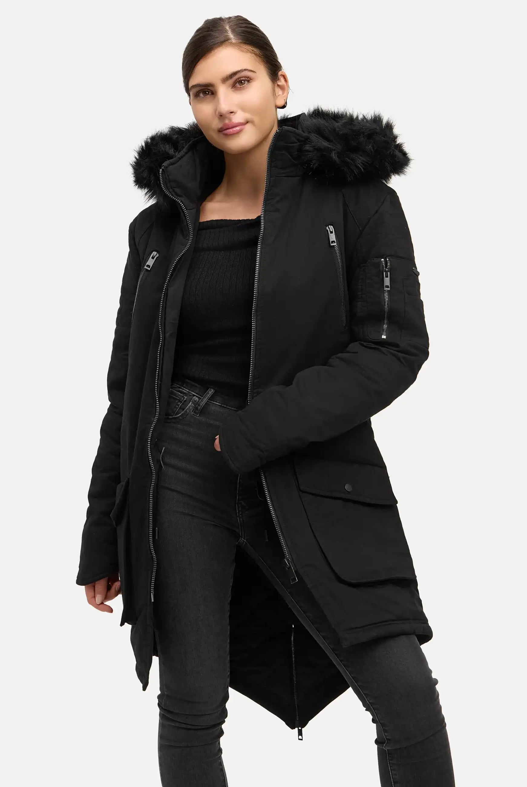 Eine Frau trägt den trueprodigy Harvey F Damenparka mit Kapuze und Kunstfell in Schwarz, gepaart mit einem schwarzen Pullover und einer schmal geschnittenen Jeans vor einem schlichten hellen Hintergrund. Sie steht mit einer Hand in der Tasche und zeigt einen neutralen Gesichtsausdruck.