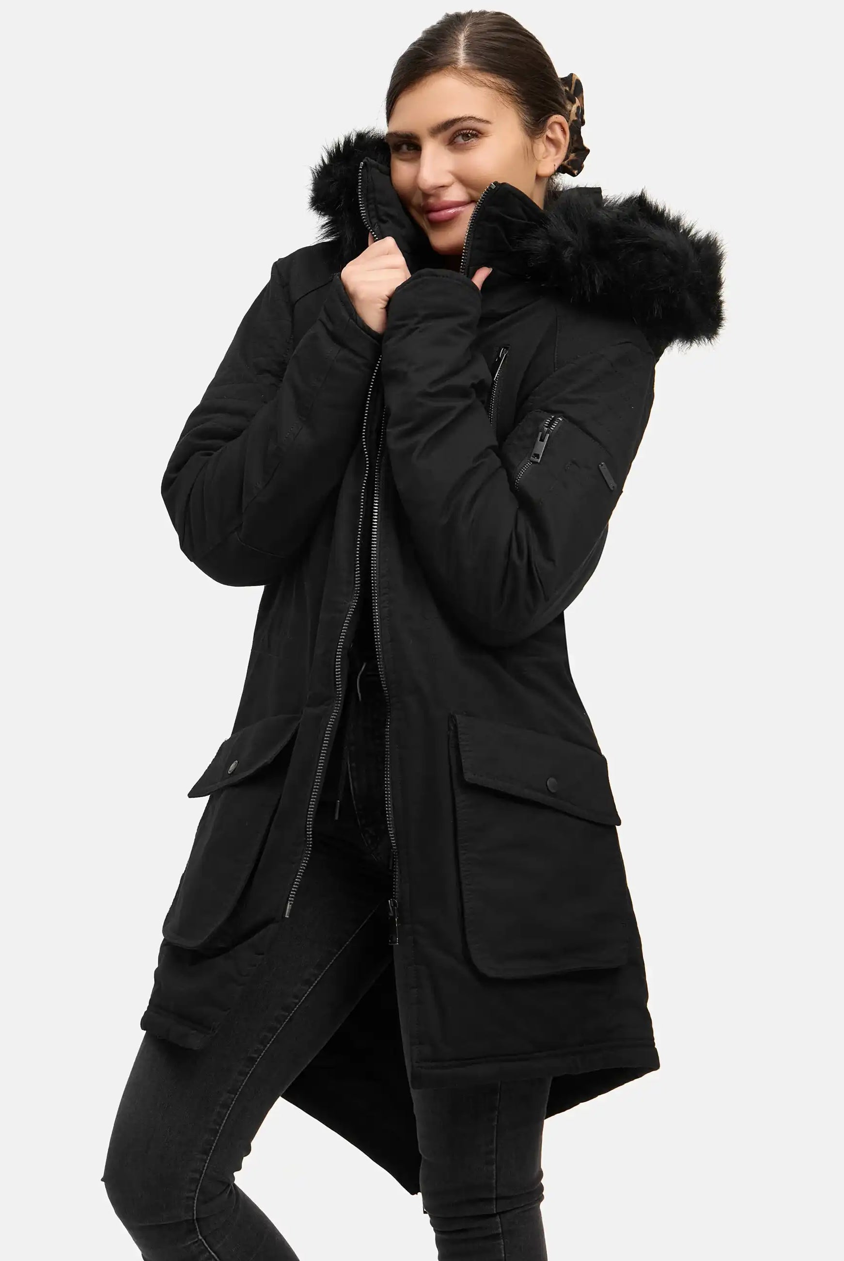 Eine Person steht vor einem schlichten Hintergrund und trägt den trueprodigy Harvey F Damenparka mit Kapuze und Kunstfell, wobei sie das Halsband eng an ihr Gesicht hält und leicht lächelt.