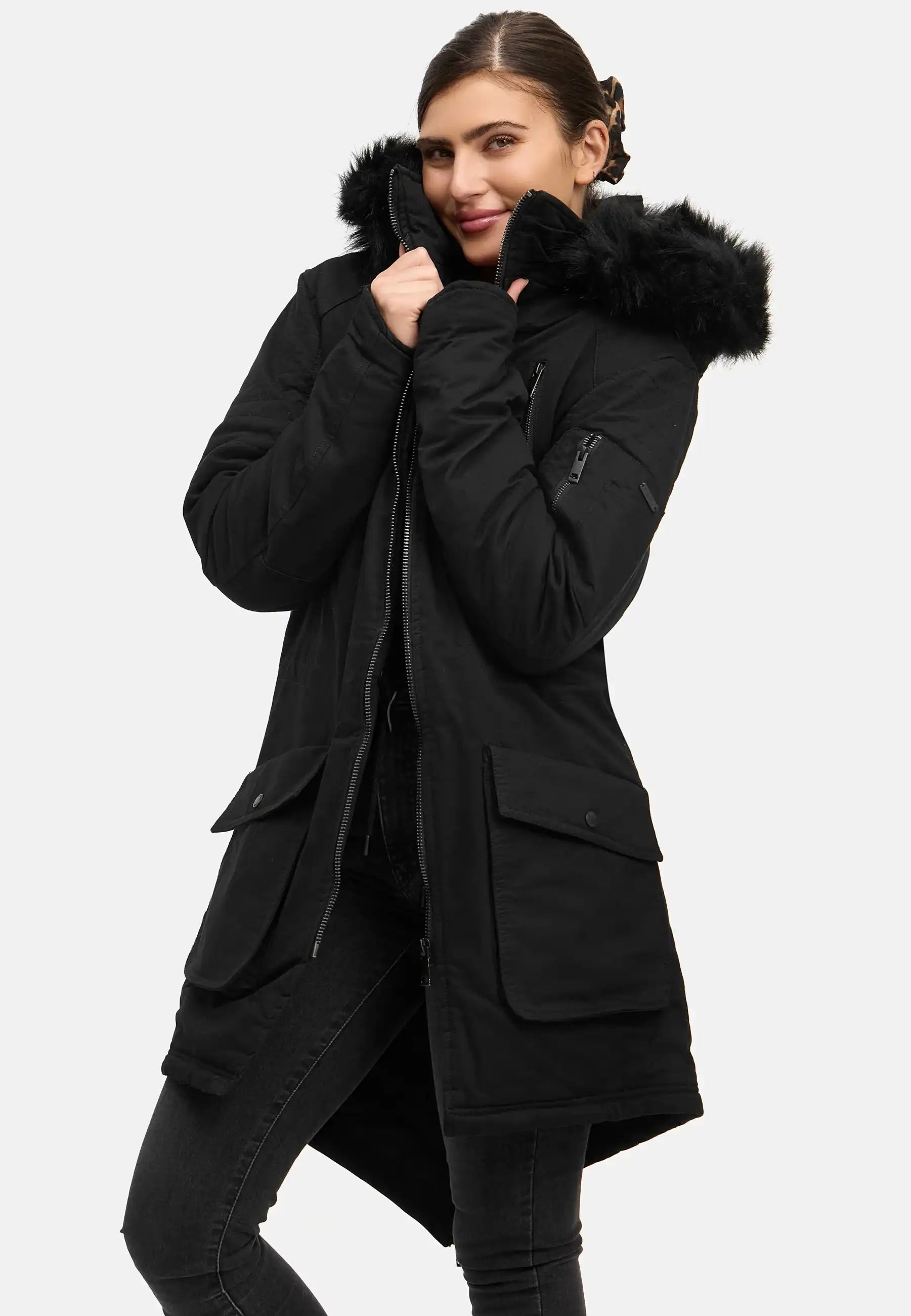 Eine Person steht vor einem schlichten Hintergrund und trägt den trueprodigy Harvey F Damenparka mit Kapuze und Kunstfell, wobei sie das Halsband eng an ihr Gesicht hält und leicht lächelt.