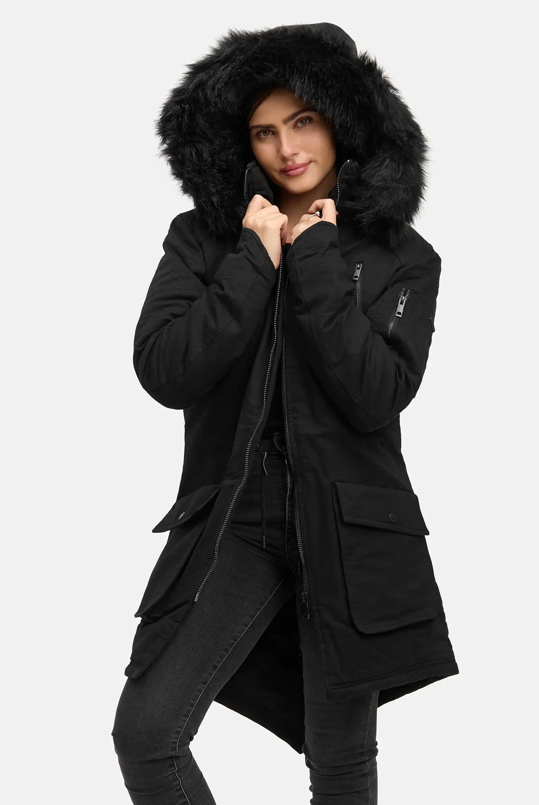 Eine Person in einem langen schwarzen trueprodigy Harvey F Damenparka mit Kapuze und Kunstfell, dunkle Hose, hält die Kapuze an ihr Gesicht. Der Hintergrund ist einfarbig und hell.