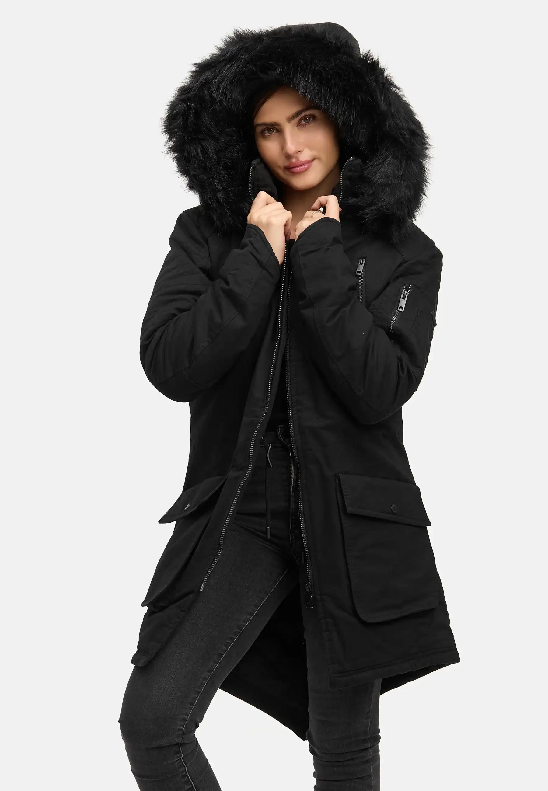 Eine Person in einem langen schwarzen trueprodigy Harvey F Damenparka mit Kapuze und Kunstfell, dunkle Hose, hält die Kapuze an ihr Gesicht. Der Hintergrund ist einfarbig und hell.