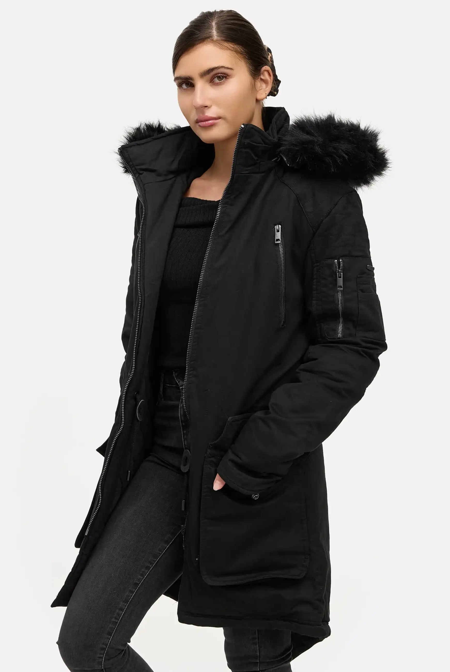 Eine Person trägt den trueprodigy Harvey F Damenparka mit Kapuze und Kunstfell in Schwarz, gepaart mit einem schwarzen Oberteil und einer dunklen Jeans, steht vor einem schlichten hellen Hintergrund und schaut mit neutralem Blick in die Kamera.