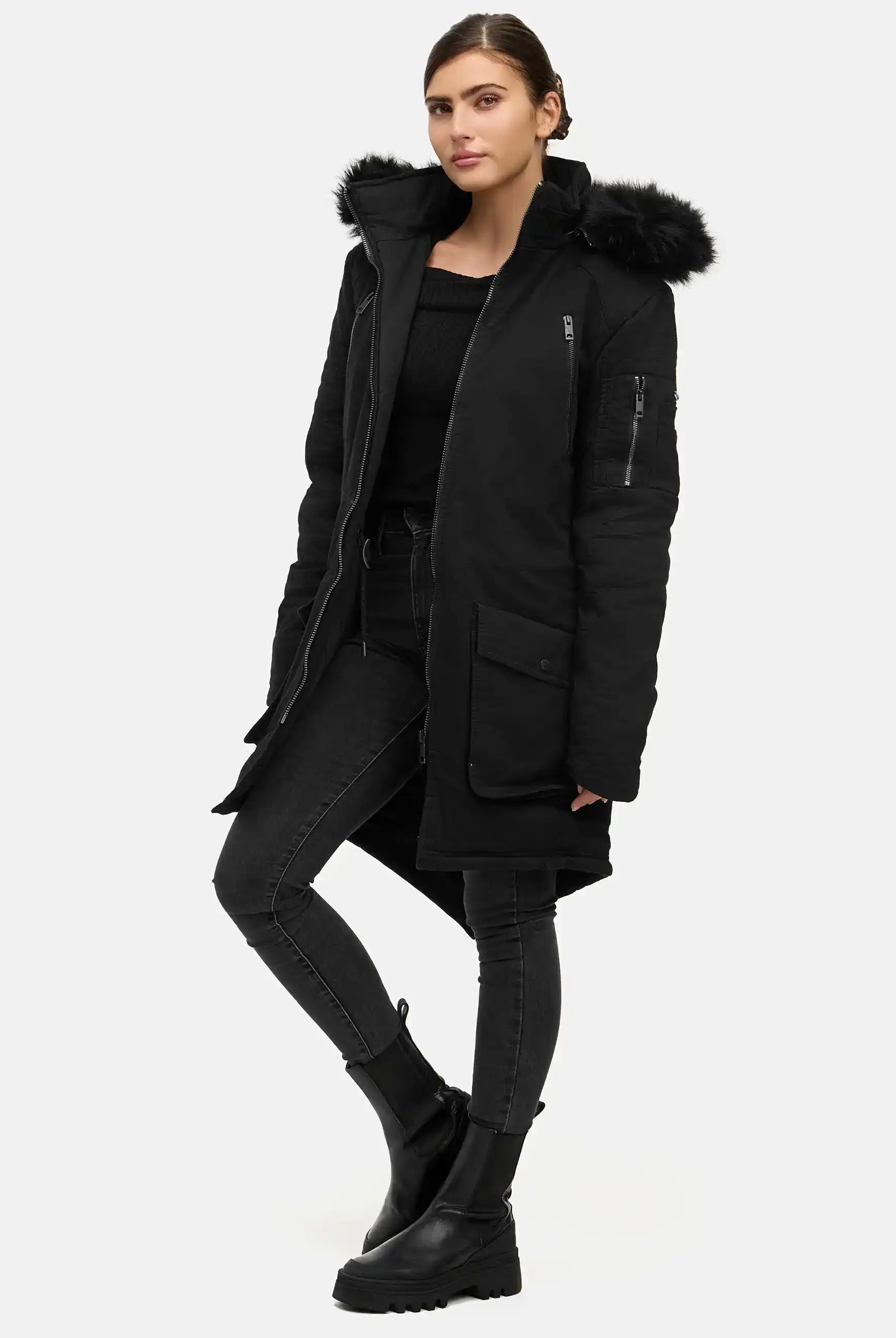 Eine Person steht mit dem Gesicht nach vorne in einem schwarzen trueprodigy Harvey F Damenparka mit Kapuze und Kunstfell, slim-fit gestylt, mit einem schwarzen Pullover, Skinny Jeans und Plateaustiefeln vor einem schlichten hellen Hintergrund.