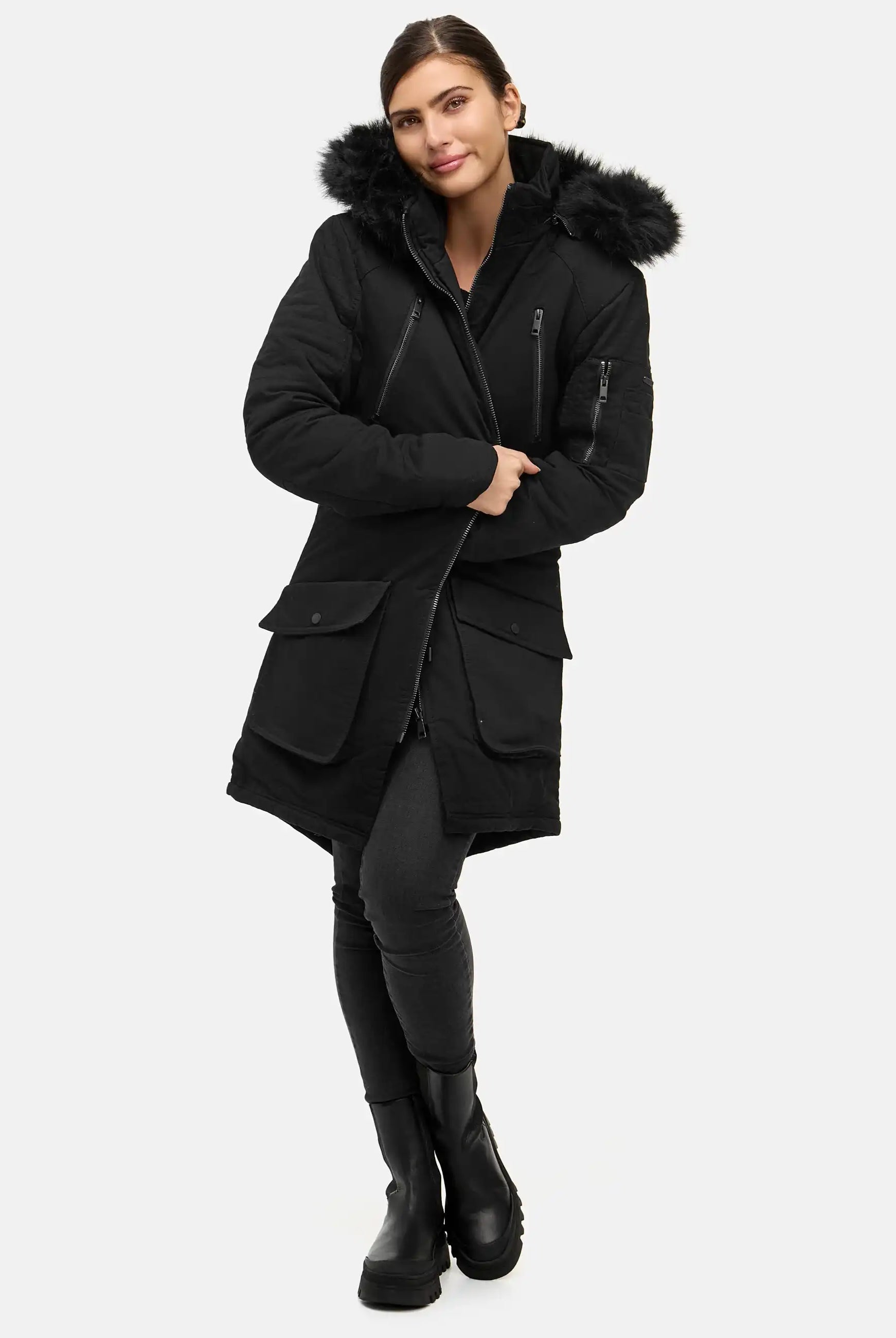 Eine Frau posiert vor einem schlichten Hintergrund in einem originalgetreuen Harvey F Damenparka mit Kapuze und Kunstfell, gepaart mit dunklen Leggings und schwarzen Stiefeln. Sie kreuzt ein Bein vor dem anderen und lächelt leicht.