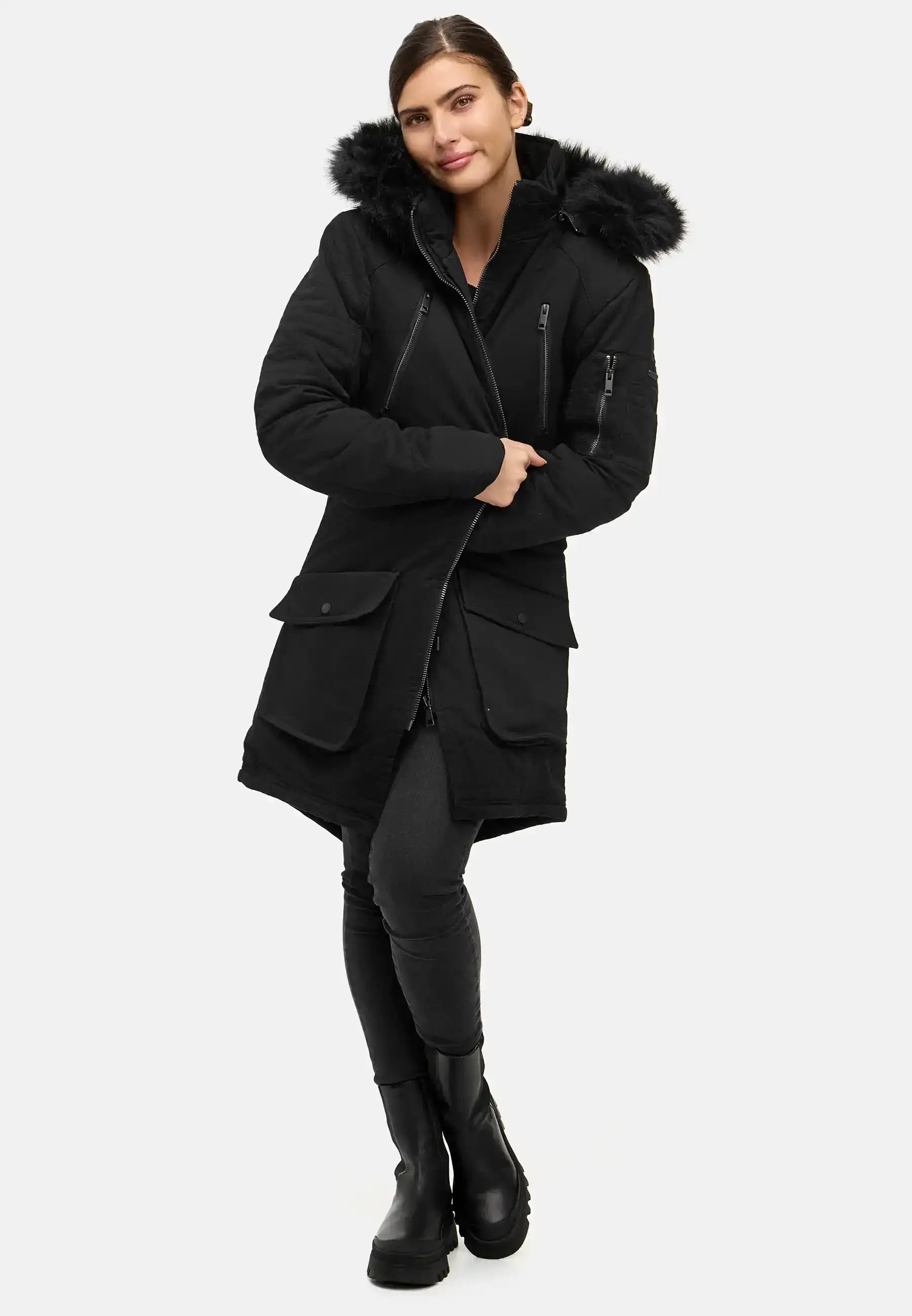 Eine Frau posiert vor einem schlichten Hintergrund in einem originalgetreuen Harvey F Damenparka mit Kapuze und Kunstfell, gepaart mit dunklen Leggings und schwarzen Stiefeln. Sie kreuzt ein Bein vor dem anderen und lächelt leicht.