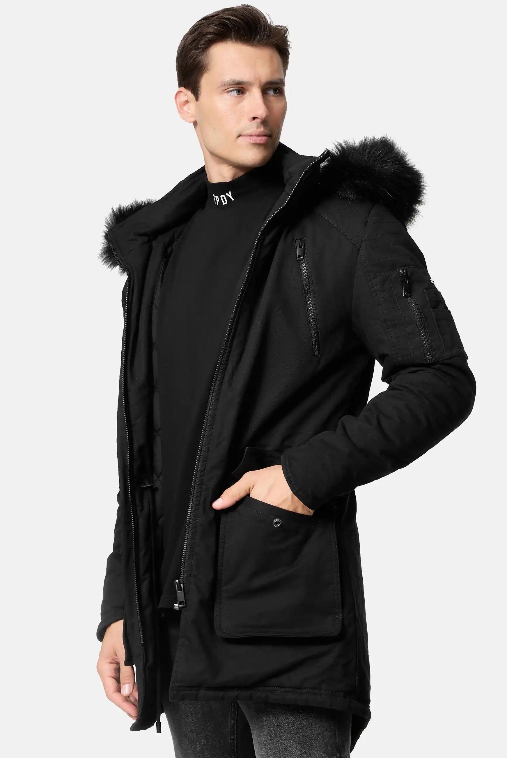 Ein Mann, der den trueprodigy Harvey Parka mit Kapuze und Kunstfell mit abnehmbarer Kapuze und geräumigen Fronttaschen trägt, steht leicht zur Seite gedreht und schaut vor einem schlichten hellen Hintergrund von der Kamera weg.