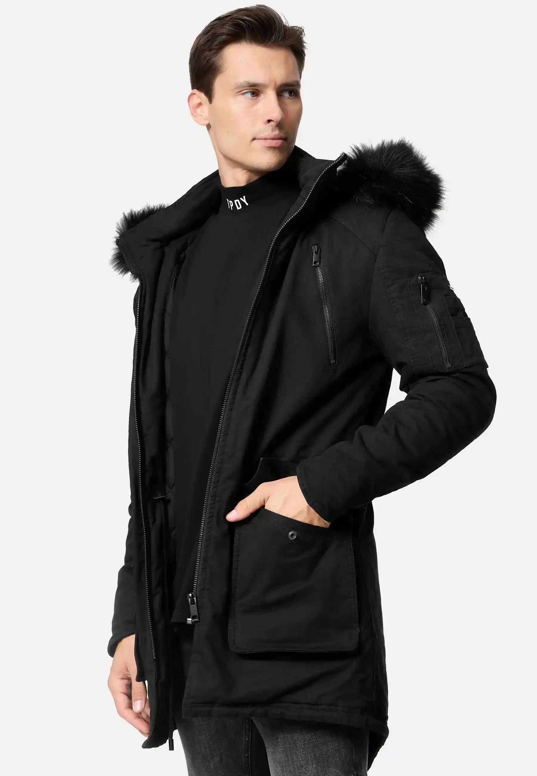 Ein Mann, der den trueprodigy Harvey Parka mit Kapuze und Kunstfell mit abnehmbarer Kapuze und geräumigen Fronttaschen trägt, steht leicht zur Seite gedreht und schaut vor einem schlichten hellen Hintergrund von der Kamera weg.