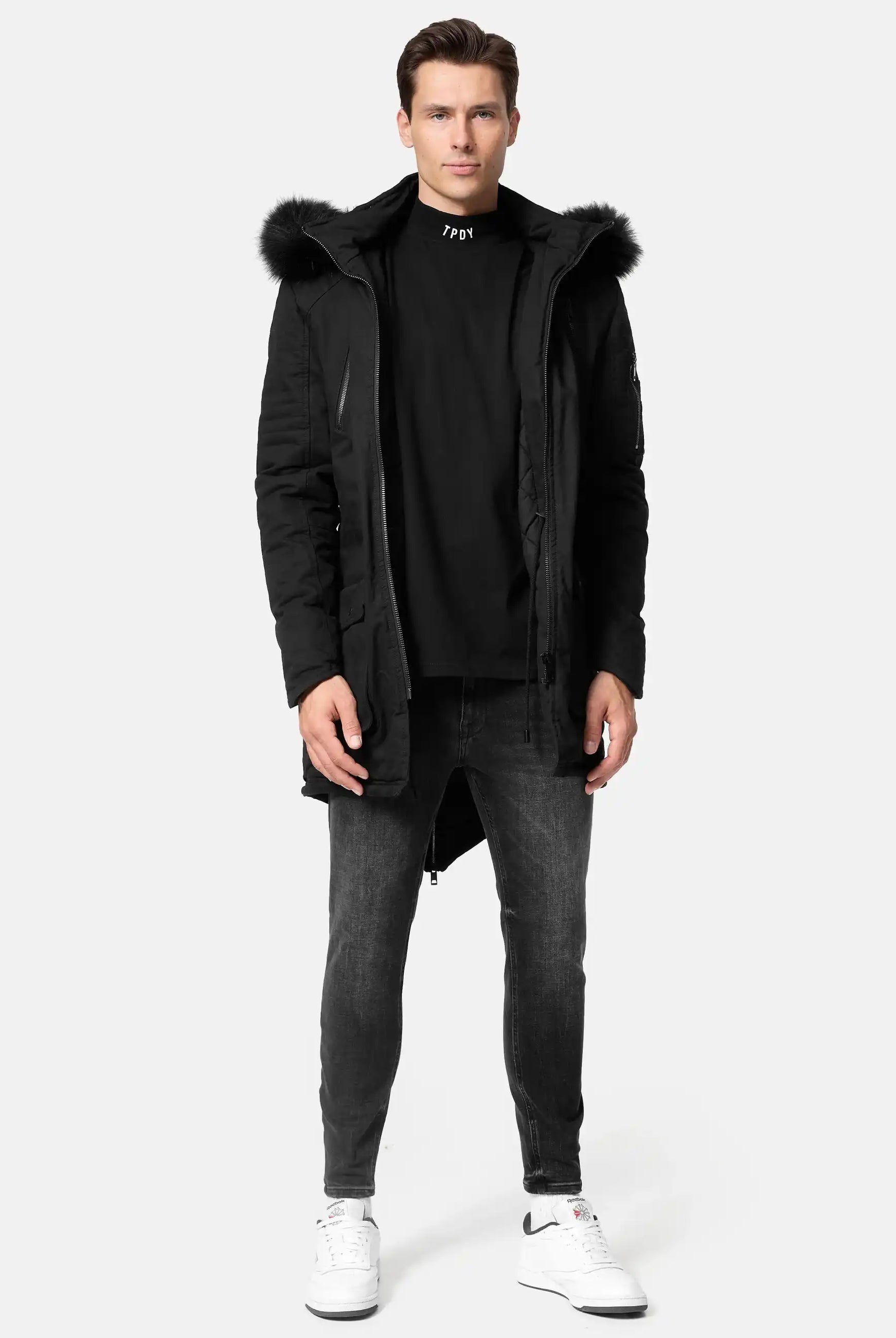 Ein Mann steht mit dem Blick nach vorne und trägt den trueprodigy Harvey Parka mit Kapuze und Kunstfell, gepaart mit einem schwarzen Rollkragenpullover, Slim-Fit-Jeans und weißen Nike-Sneakers vor einem schlichten hellgrauen Hintergrund.