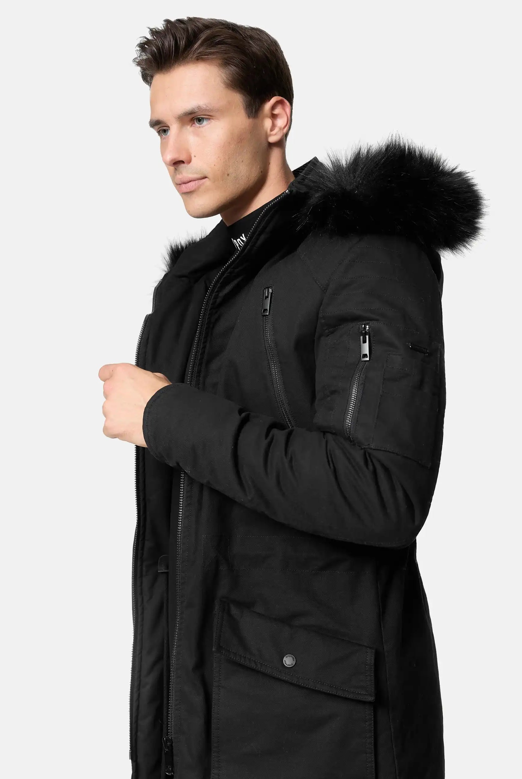 Ein Mann trägt den trueprodigy Harvey Parka mit Kapuze und Kunstfell in Slim-Fit, mit schwarzer Kapuze mit Kunstfellbesatz, vor einem hellen Hintergrund, leicht zur Seite gedreht und eine Hand am Reißverschluss.