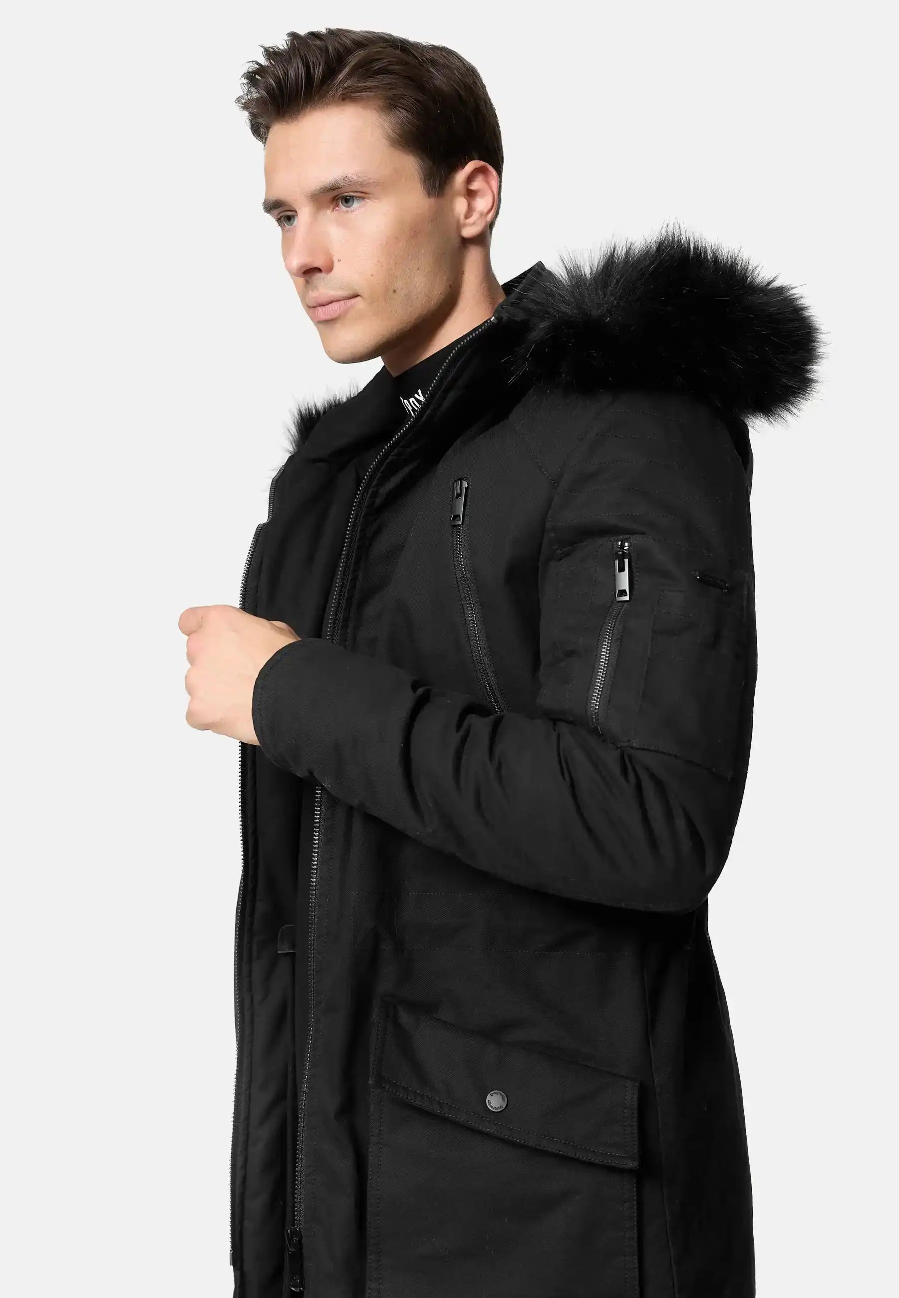 Ein Mann trägt den trueprodigy Harvey Parka mit Kapuze und Kunstfell in Slim-Fit, mit schwarzer Kapuze mit Kunstfellbesatz, vor einem hellen Hintergrund, leicht zur Seite gedreht und eine Hand am Reißverschluss.