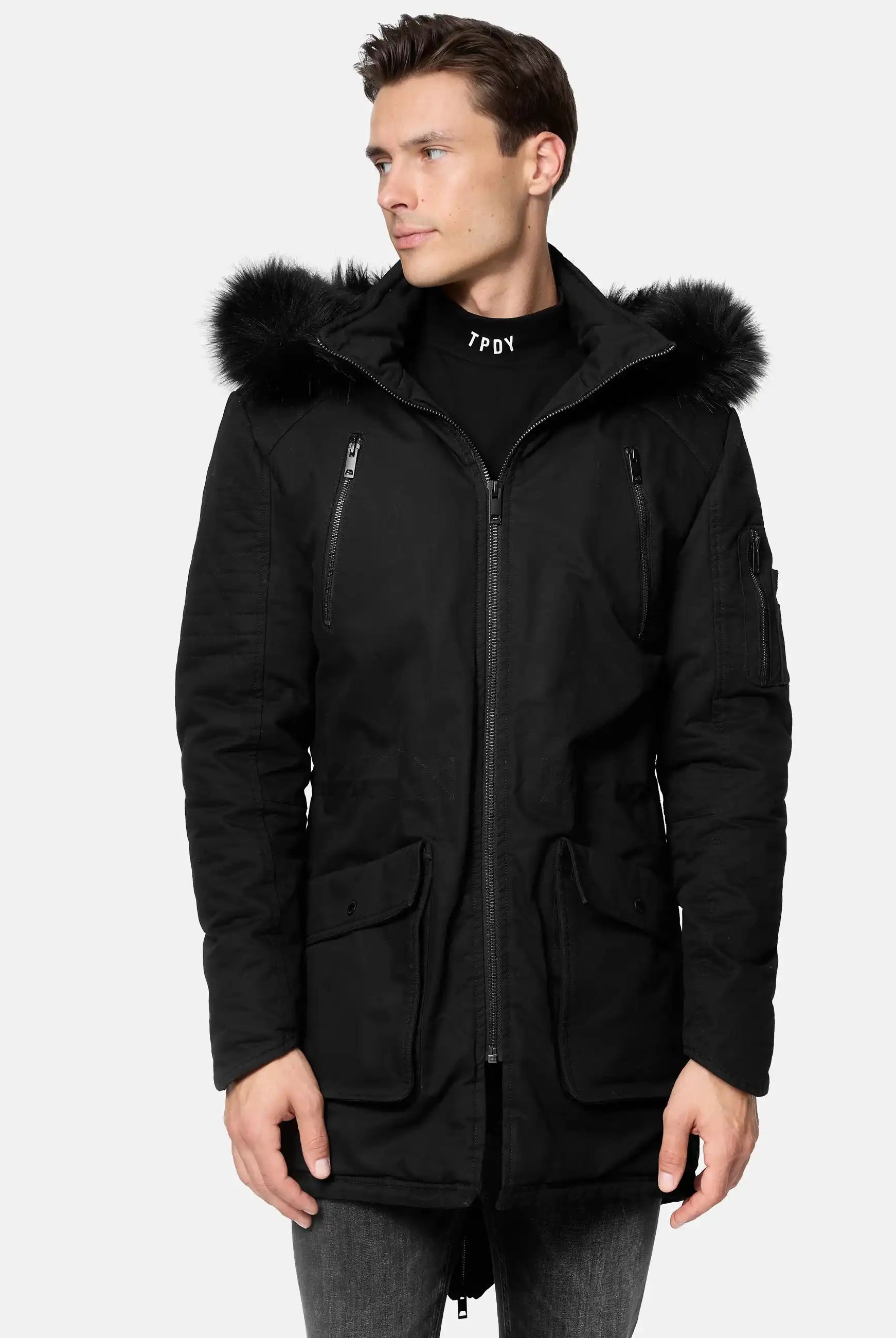 Ein Mann trägt den trueprodigy Harvey Parka mit Kapuze und Kunstfell, der mit einer pelzgefütterten, abnehmbaren Kapuze, Brusttaschen mit Reißverschluss und Pattentaschen an der Taille ausgestattet ist. Er steht leicht nach links und kombiniert ihn mit einer grauen Jeans.