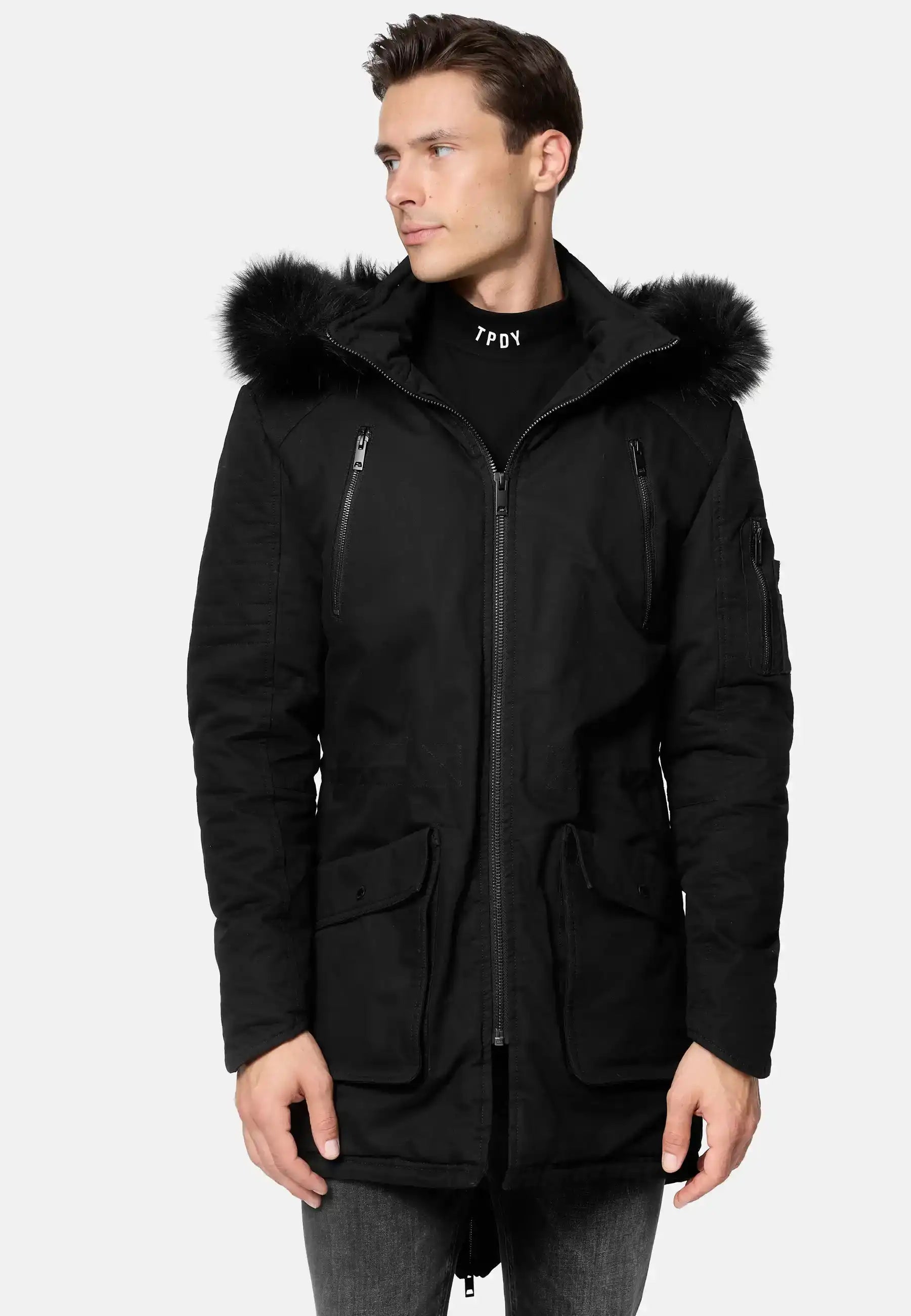 Ein Mann trägt den trueprodigy Harvey Parka mit Kapuze und Kunstfell, der mit einer pelzgefütterten, abnehmbaren Kapuze, Brusttaschen mit Reißverschluss und Pattentaschen an der Taille ausgestattet ist. Er steht leicht nach links und kombiniert ihn mit einer grauen Jeans.