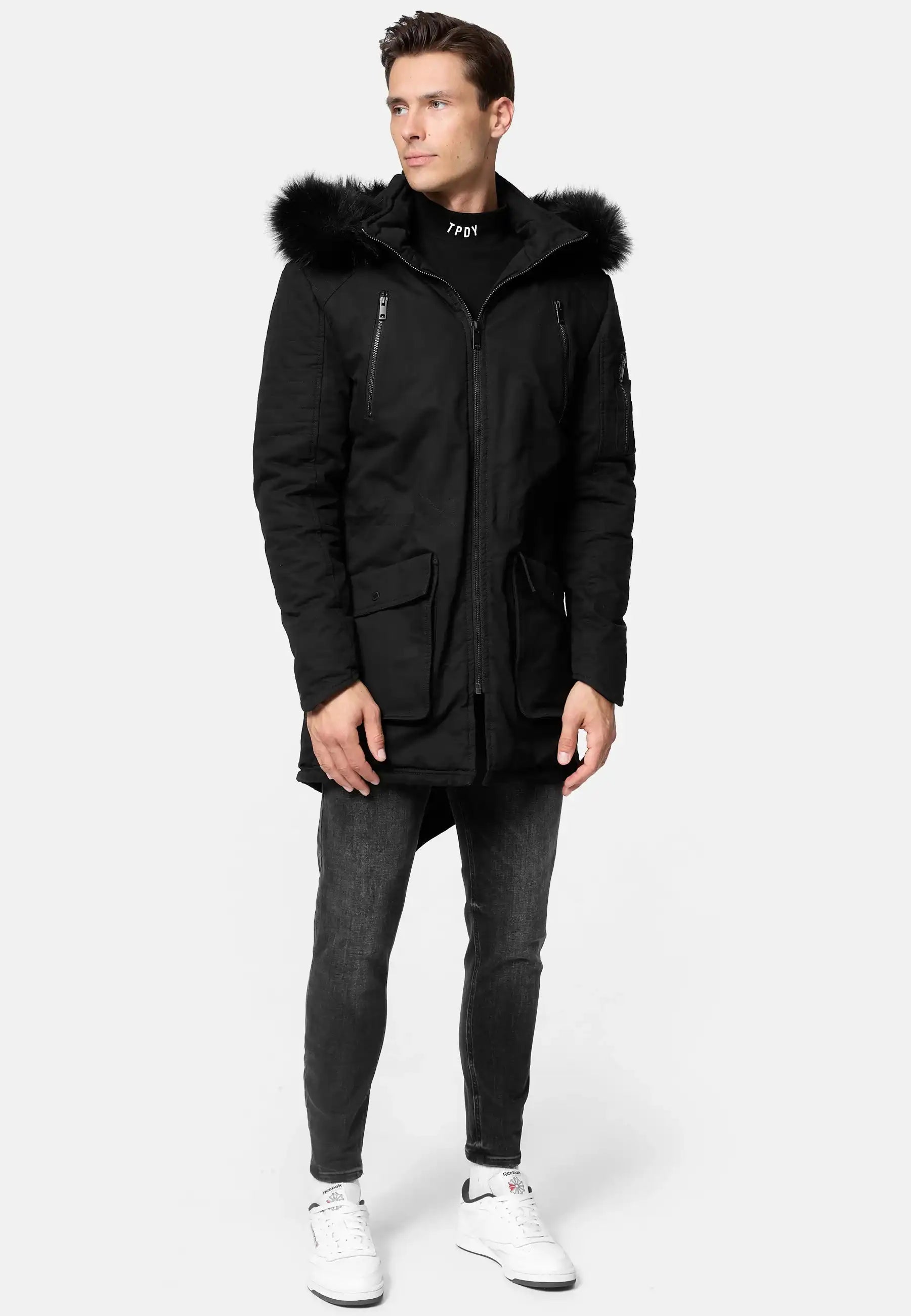 Ein Mann steht seitlich und trägt den trueprodigy Harvey Parka mit Kapuze und Kunstfell in Schwarz, eine dunkle Slim-Fit-Jacke sowie weiße Nike Sneakers. Der Hintergrund ist schlicht und hellgrau.