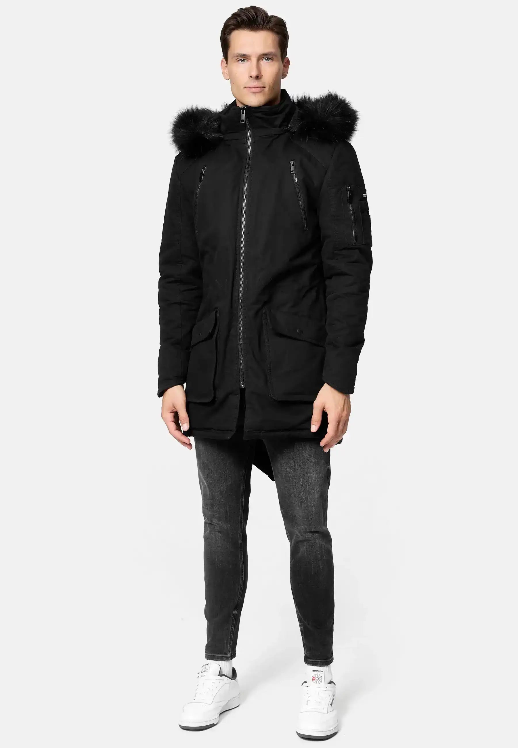 Ein Mann steht mit dem Blick nach vorne und trägt den schwarzen Harvey Parka mit Kapuze und Kunstfell von trueprodigy, schmale dunkle Jeans und weiße Turnschuhe vor einem schlichten hellgrauen Hintergrund.