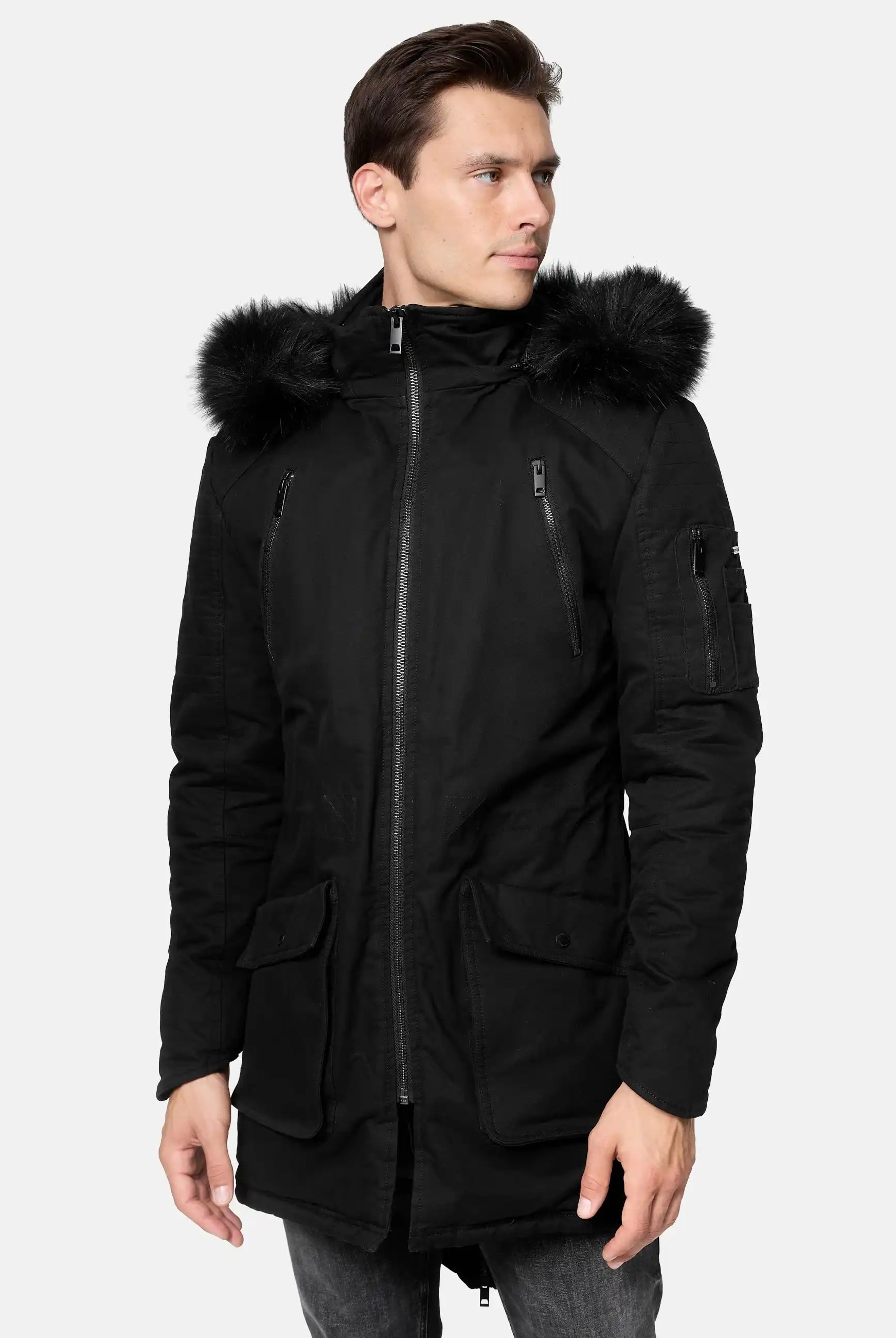 Ein Mann in einem schwarzen Harvey Parka mit Kapuze und Kunstfell von trueprodigy, mit abnehmbarem Kapuze und mehreren Taschen, steht leicht gedreht vor einem schlichten hellen Hintergrund.