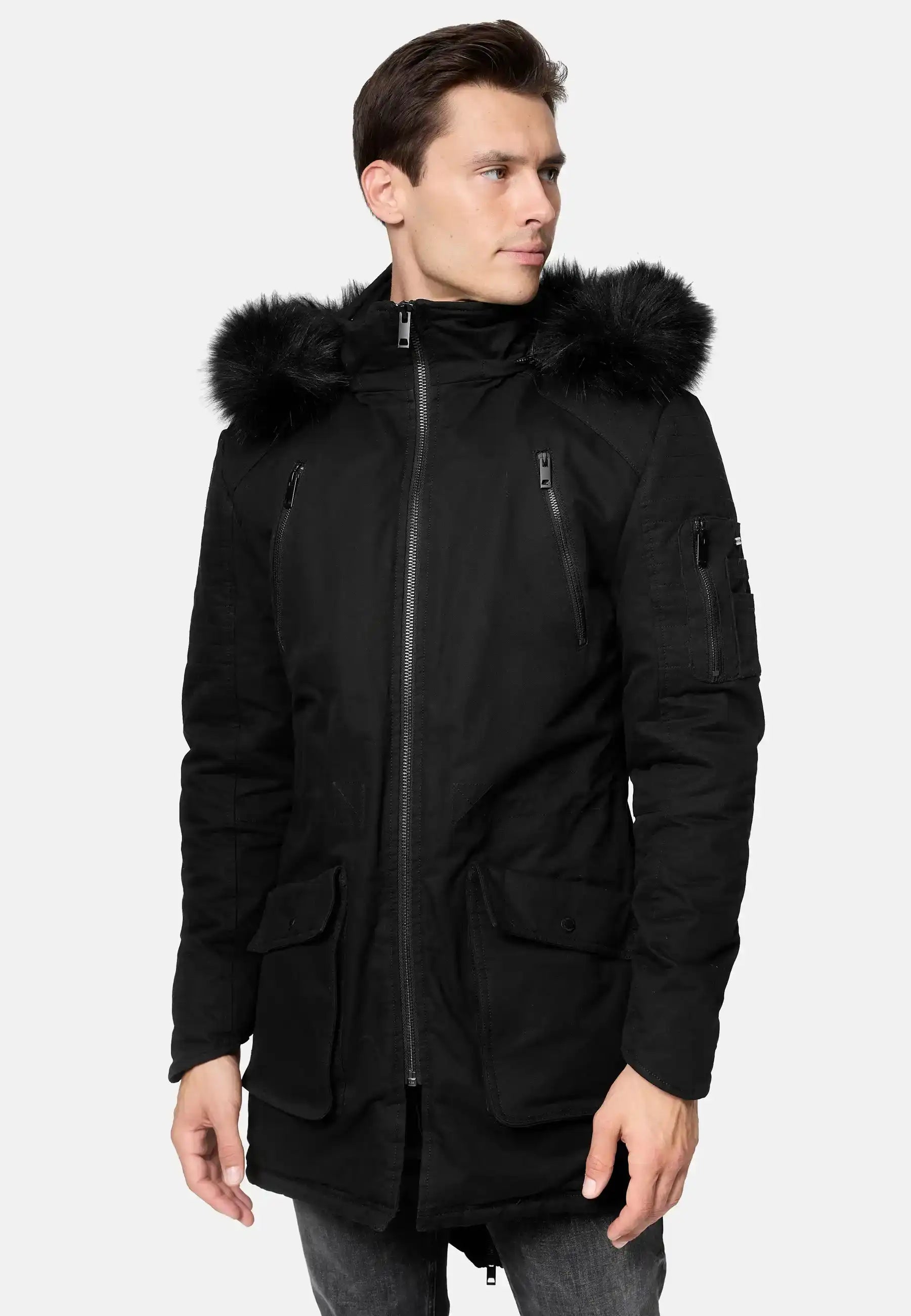 Ein Mann in einem schwarzen Harvey Parka mit Kapuze und Kunstfell von trueprodigy, mit abnehmbarem Kapuze und mehreren Taschen, steht leicht gedreht vor einem schlichten hellen Hintergrund.