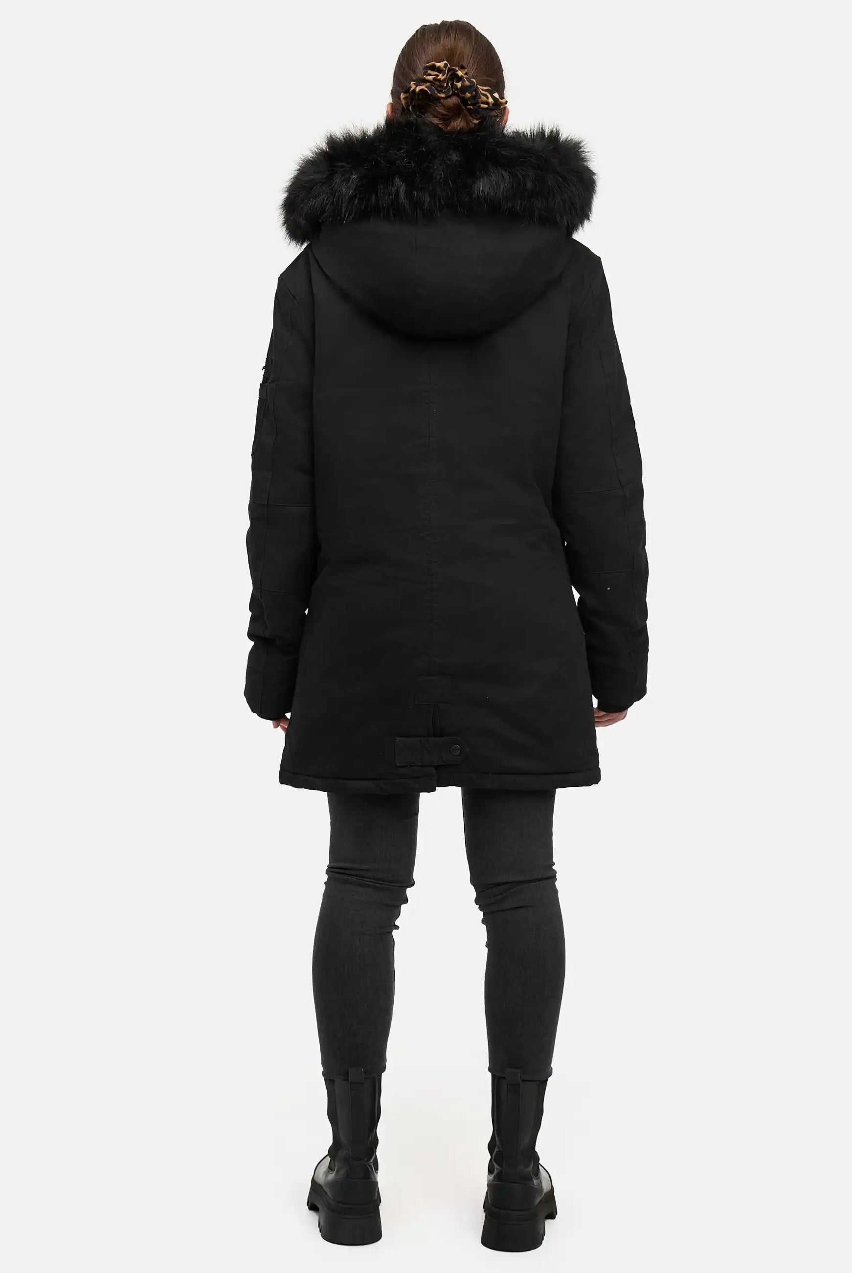 Eine Person steht mit dem Gesicht zur Wand und trägt den trueprodigy Julius F Parka mit Kapuze und Kunstfell, gepaart mit einer schwarzen Hose und Stiefeln. Die Haare sind hochgesteckt und ein schlichter weißer Hintergrund unterstreicht die lässige Passform des Parkas.