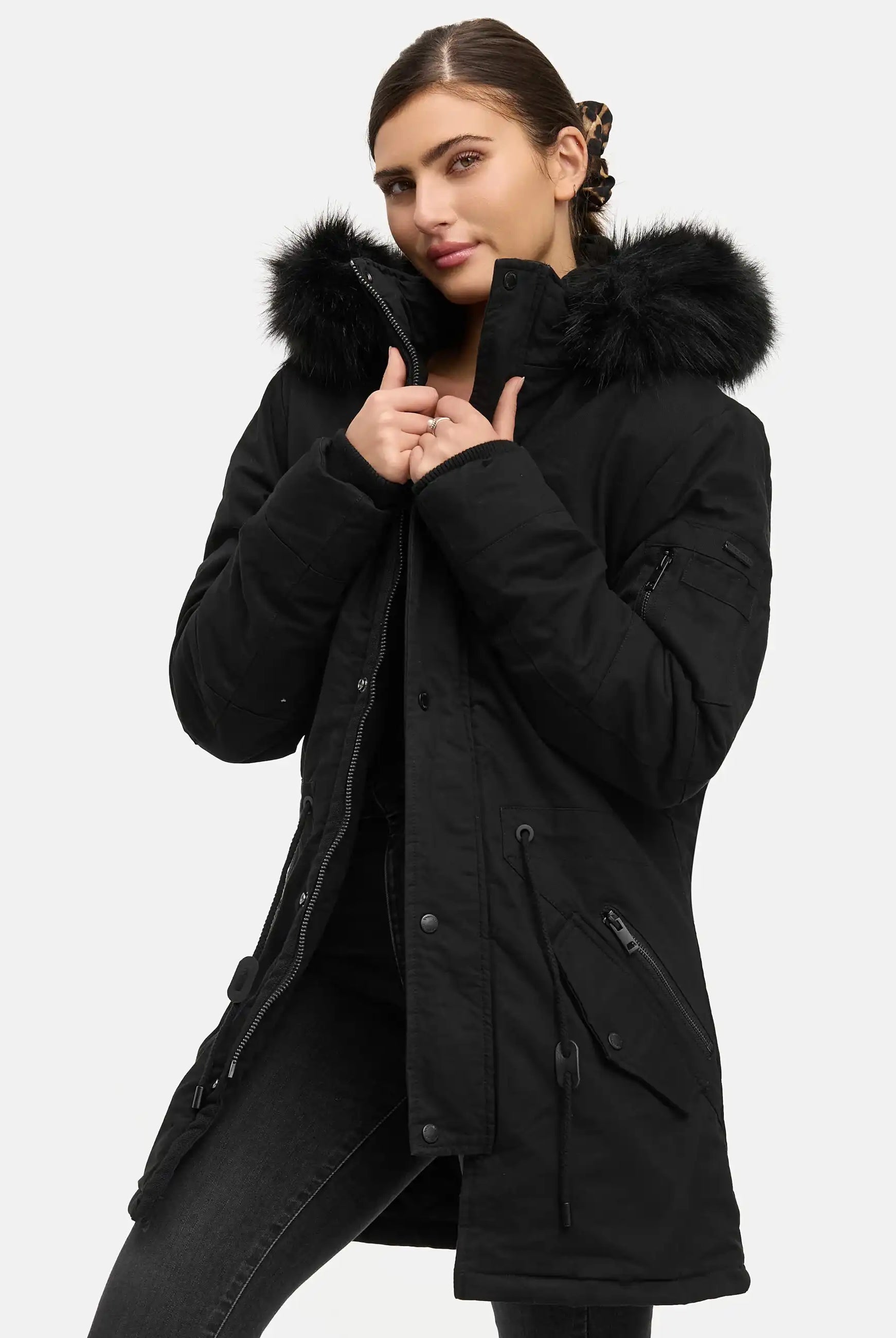 Eine Frau trägt den trueprodigy Julius F Parka mit Kapuze und Kunstfell in Schwarz, schaut in die Kamera und hält den Kragen geschlossen. Der lange Parka ist mit mehreren Taschen und Reißverschlussdetails ausgestattet und wird vor einem schlichten weißen Hintergrund gezeigt.