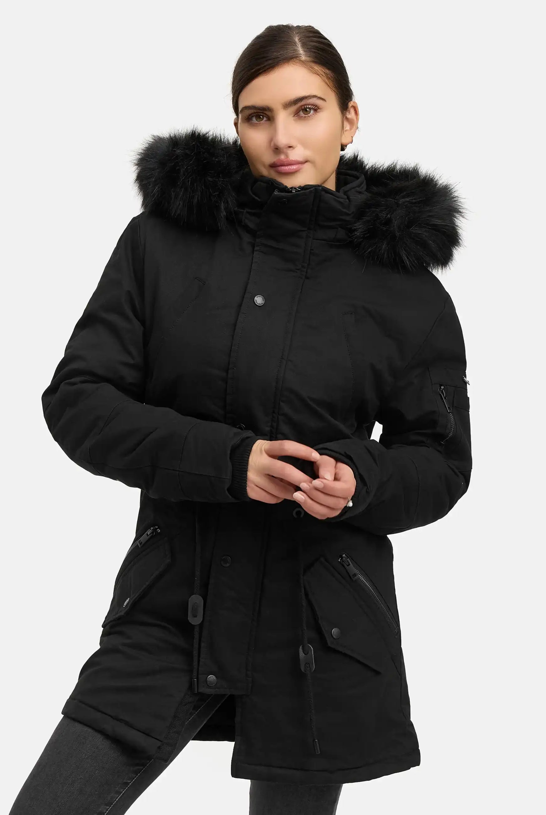 Eine Person, die den Julius F Parka mit Kapuze und Kunstfell von trueprodigy trägt, steht vor einem schlichten weißen Hintergrund und blickt mit einem leichten Lächeln und voreinander geschlagenen Händen in die Kamera.