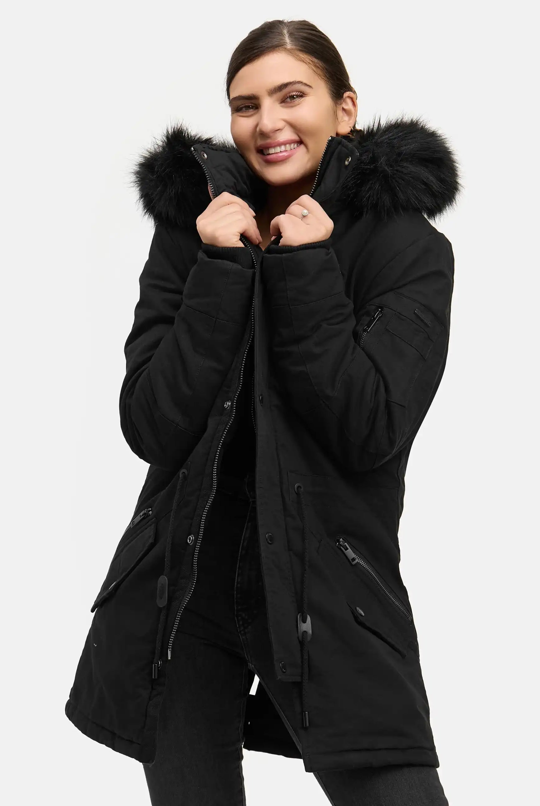 Eine Person lächelt, während sie den trueprodigy Julius F Parka mit Kapuze und Kunstfell in Schwarz trägt und den Kragen mit beiden Händen vor einem schlichten weißen Hintergrund hält.