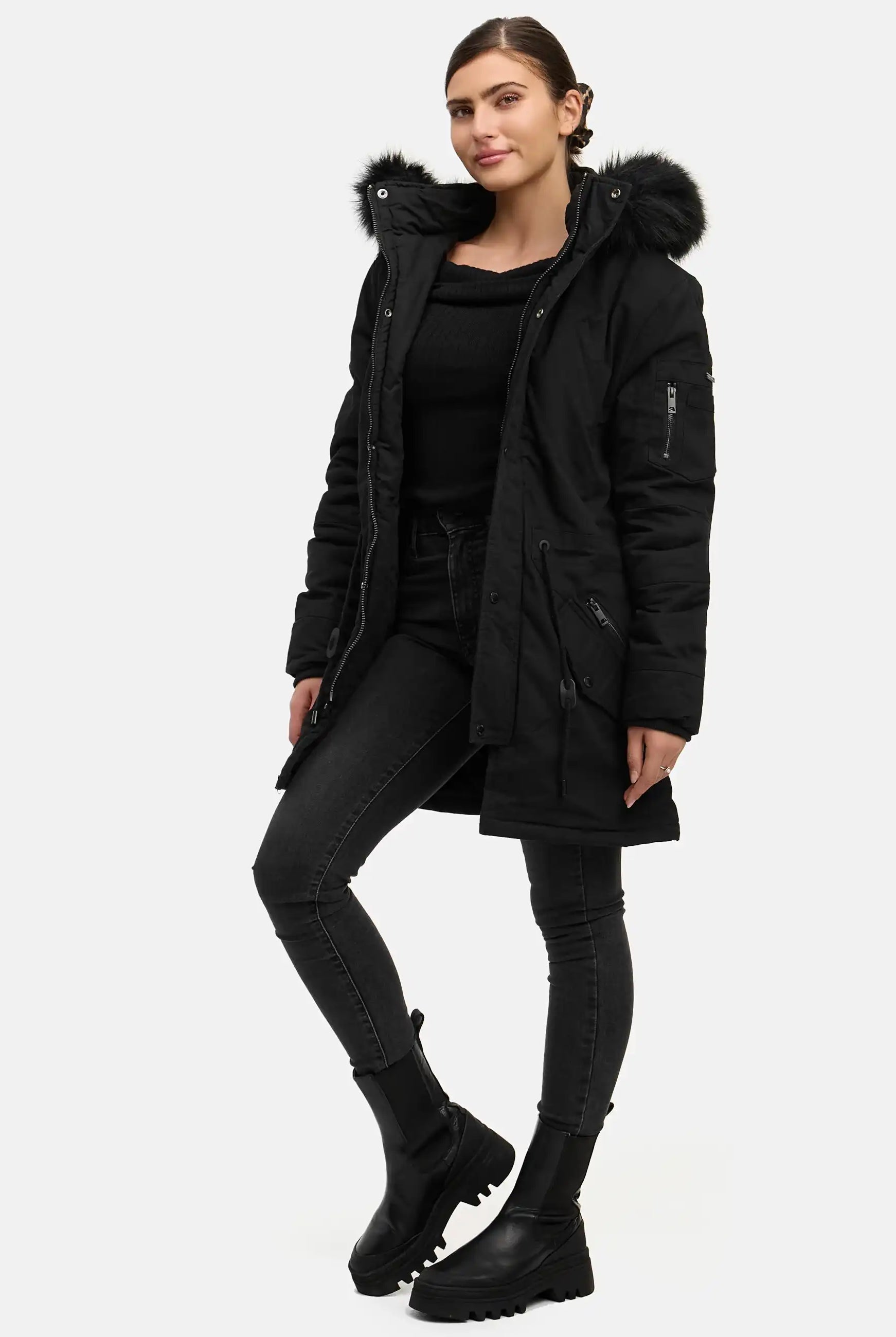 Eine Frau blickt nach vorne und trägt einen trueprodigy Julius F Parka mit Kapuze und Kunstfell, der mit einem schwarzen Oberteil, Jeans und Stiefeln gestylt ist. Eine Hand steckt in ihrer Tasche, ihr Gesichtsausdruck ist neutral, und der Hintergrund ist schlicht weiß.