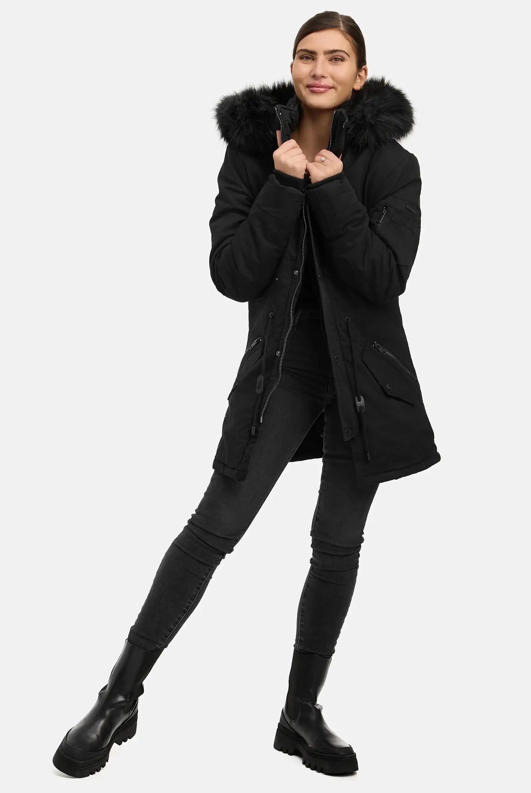 Eine Person in einem trueprodigy Julius F Parka mit Kapuze und Kunstfell, schwarzen Skinny Jeans und schwarzen Stiefeln steht auf einem Bein, lächelt und hält mit beiden Händen den Kragen der pelzgefütterten Kapuze vor einem schlichten weißen Hintergrund.