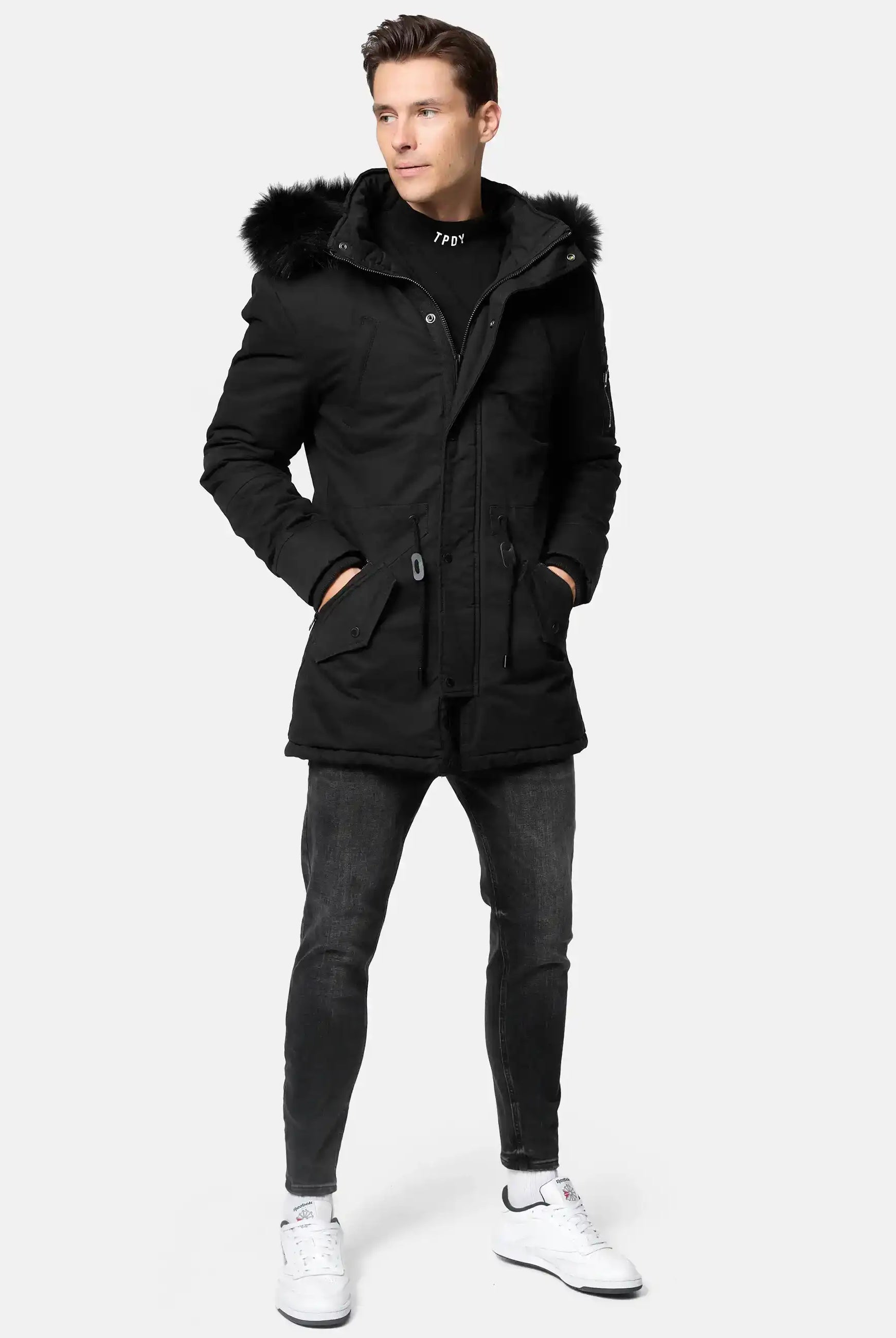 Ein Mann steht mit den Händen in den Manteltaschen und trägt den trueprodigy Julius Parka mit Kapuze und Kunstfell, gepaart mit einer schwarzen Jeans, einem schwarzen Pullover und weißen Turnschuhen vor einem schlichten hellen Hintergrund.