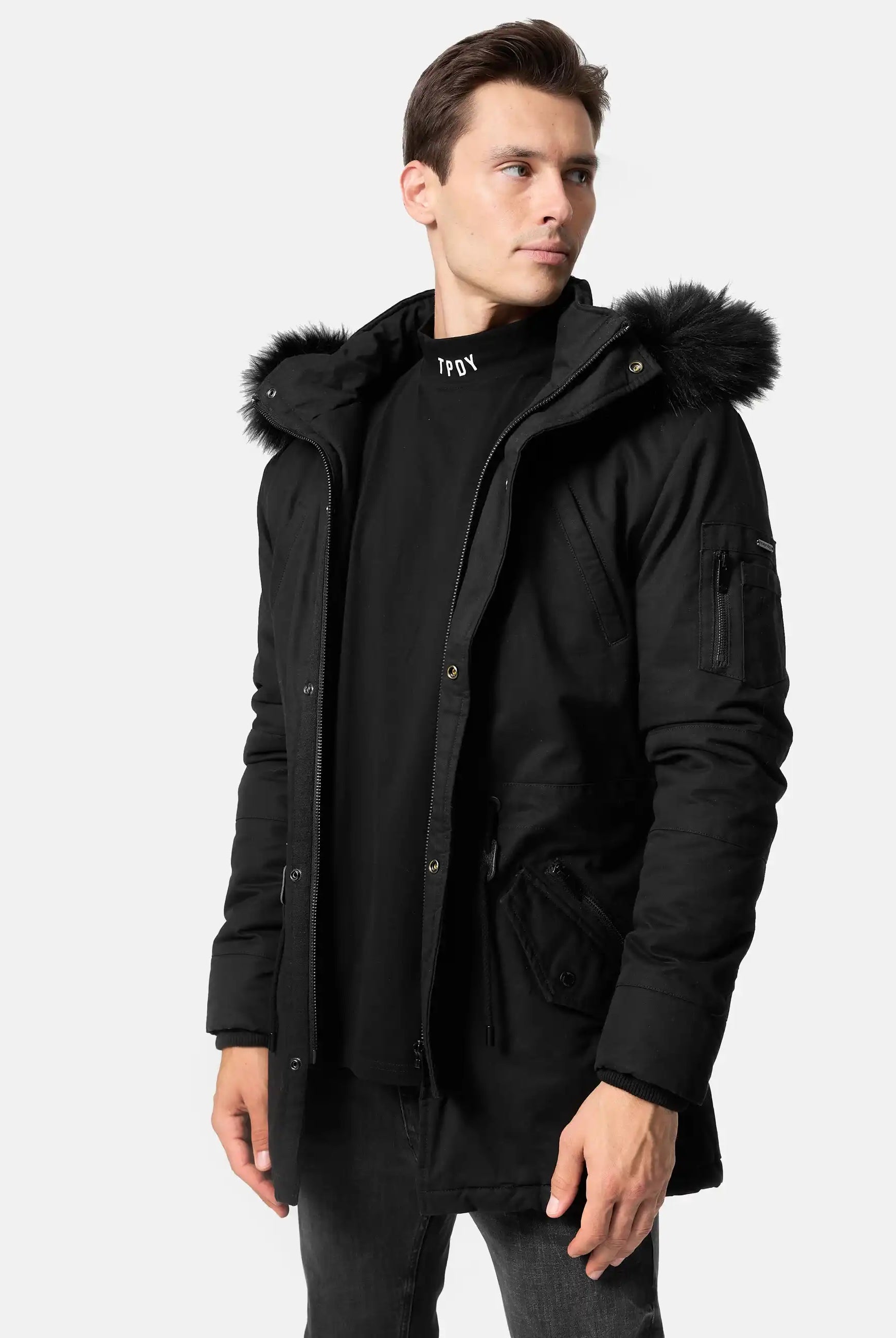 Mann trägt schwarze TRUEPRODIGY julius Winterjacke mit pelzbesetzter Kapuze und aufgesetzten Taschen; schwarzes Shirt mit „TPOY“-Aufschrift. Modernes Design, lässiger Stil, perfekt für kaltes Wetter.