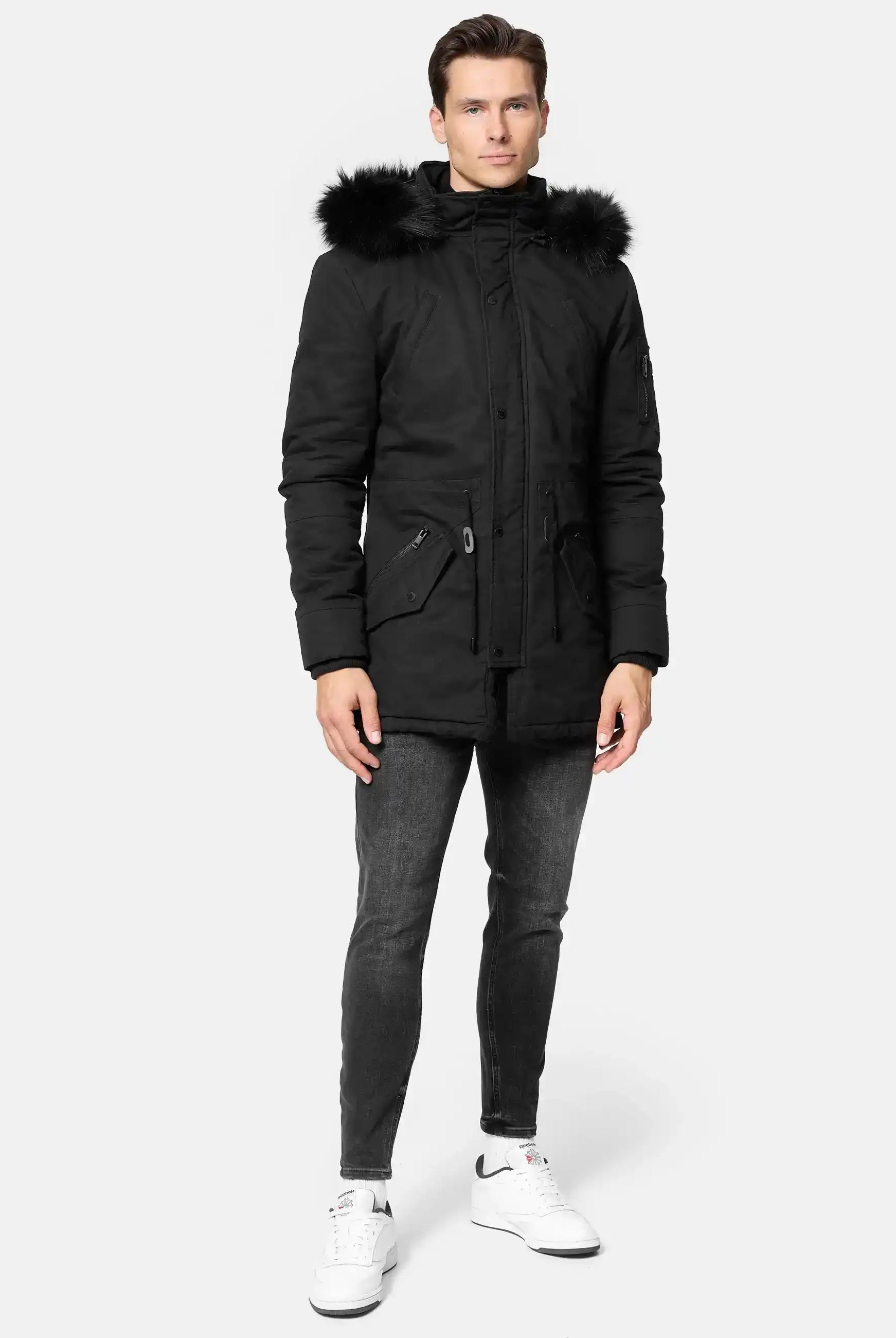 Ein Mann sieht nach vorne und trägt einen schwarzen trueprodigy Julius Parka mit Kapuze und Kunstfell, dunkelgraue Jeans und weiße Turnschuhe vor einem schlichten hellgrauen Hintergrund.