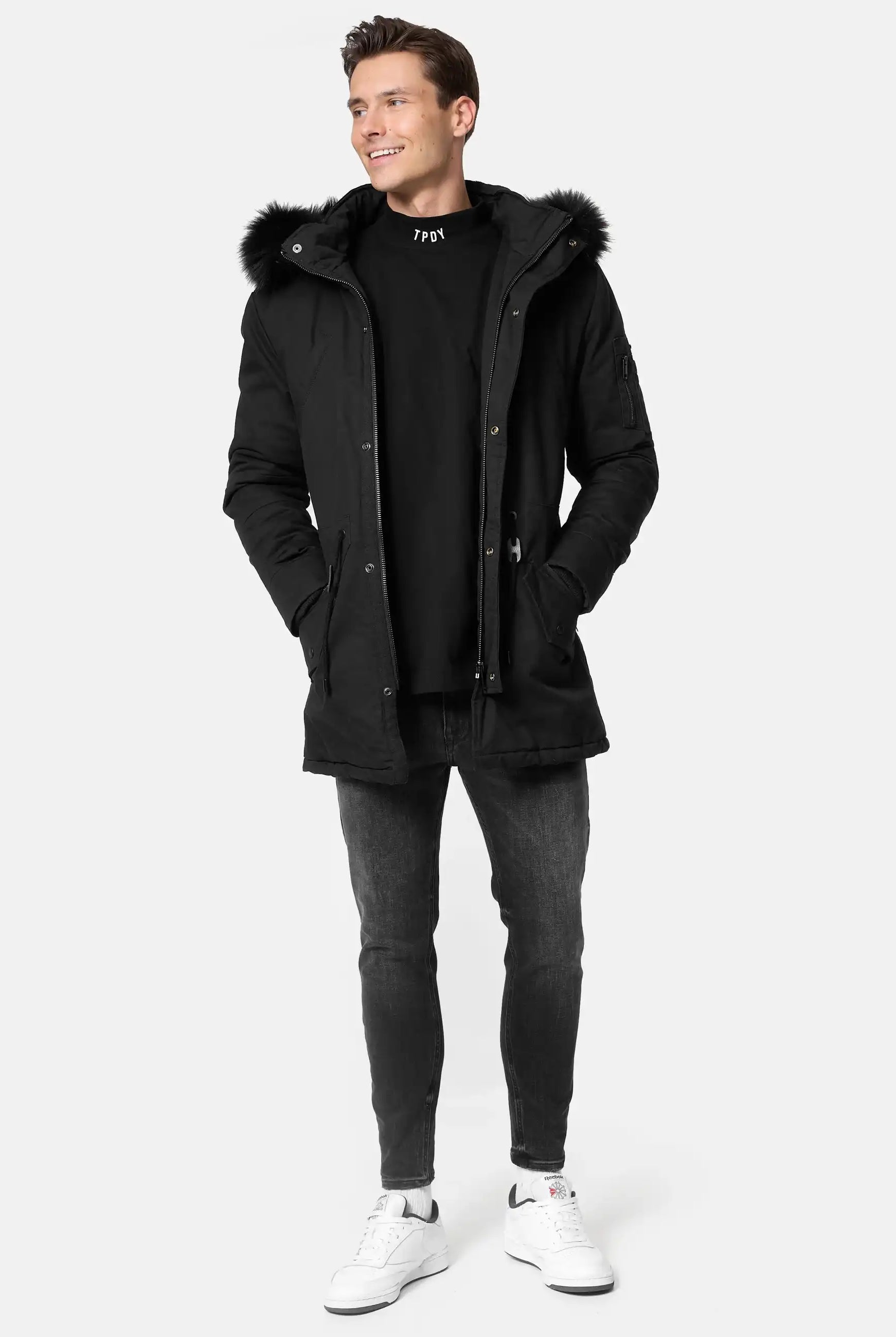 Ein Mann steht mit den Händen in den Taschen eines trueprodigy Julius Parka mit Kapuze und Kunstfell, gepaart mit einem schwarzen Sweatshirt, dunklen Skinny Jeans und weißen Turnschuhen vor einem schlichten, hellen Hintergrund.