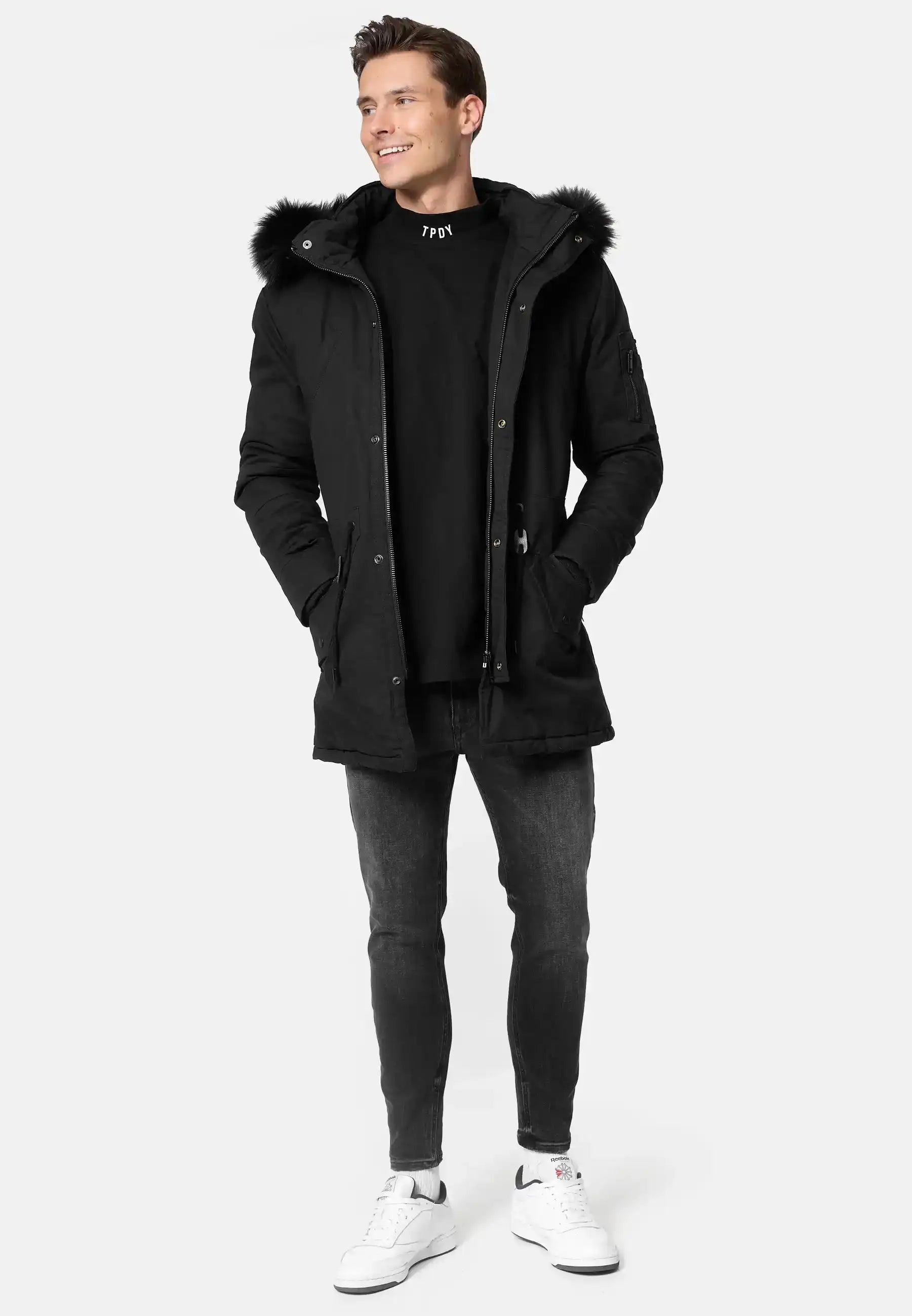 Ein Mann steht mit den Händen in den Taschen eines trueprodigy Julius Parka mit Kapuze und Kunstfell, gepaart mit einem schwarzen Sweatshirt, dunklen Skinny Jeans und weißen Turnschuhen vor einem schlichten, hellen Hintergrund.