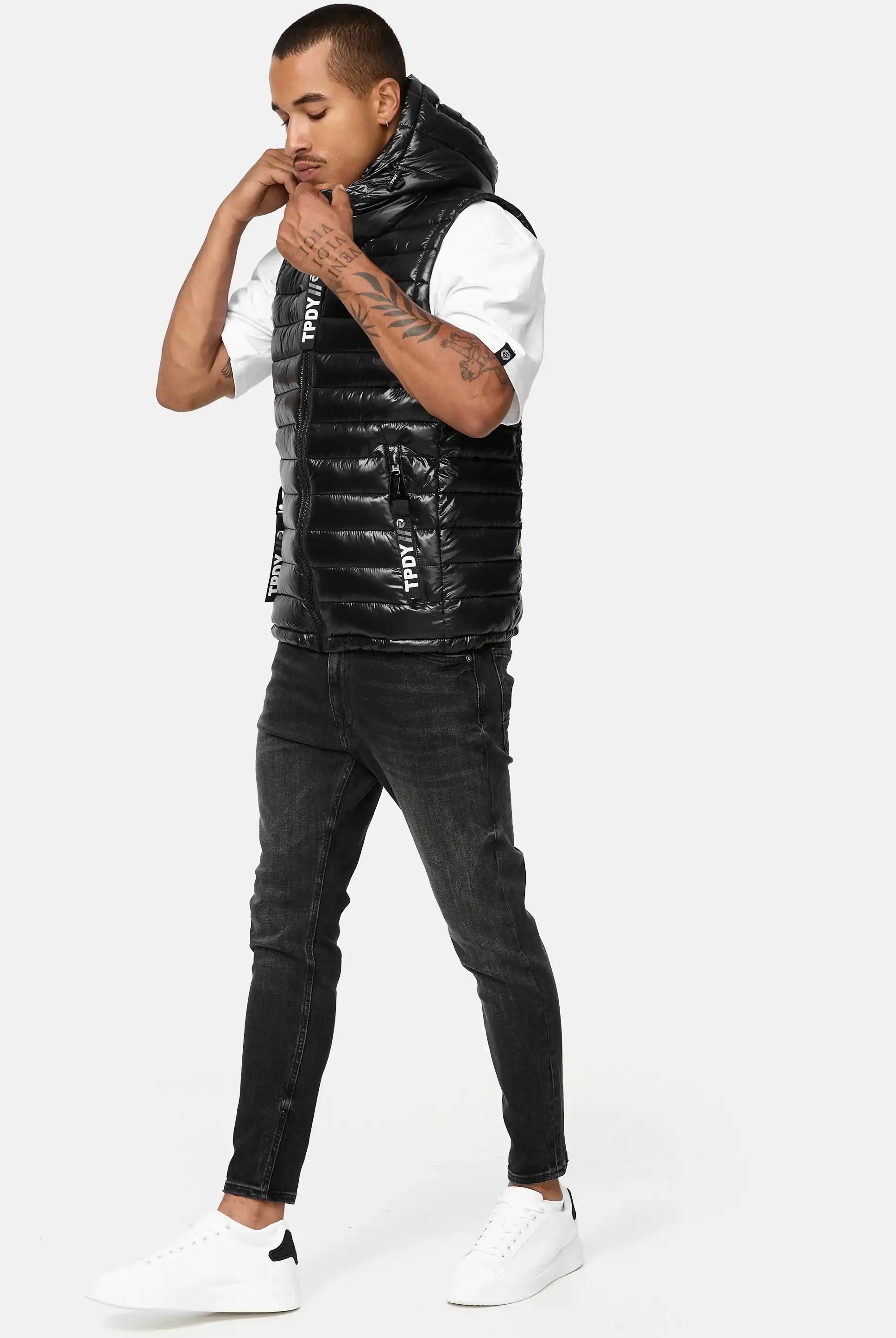 Ein Mann mit Armtattoos steht vor einem schlichten Hintergrund und blickt nach unten, während er die Kapuze seiner Trueprodigy Jared Herrenweste mit Glanz und Kapuze zurechtrückt. Er trägt ein weißes T-Shirt, schwarze Jeans und weiße Turnschuhe.