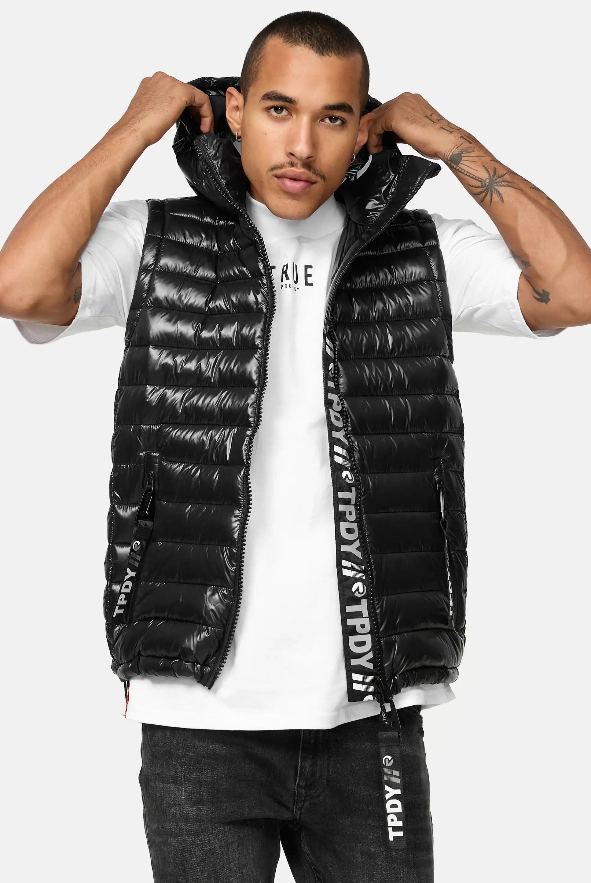 Ein Mann blickt nach vorne und trägt die trueprodigy Jared Herrenweste mit Glanz und Kapuze, eine glänzende schwarze Pufferweste mit Kapuze über einem weißen T-Shirt. Er hält die Kapuze, und die Weste ist mit horizontalen Steppungen und schwarzen Kordelzügen mit weißem Text versehen.