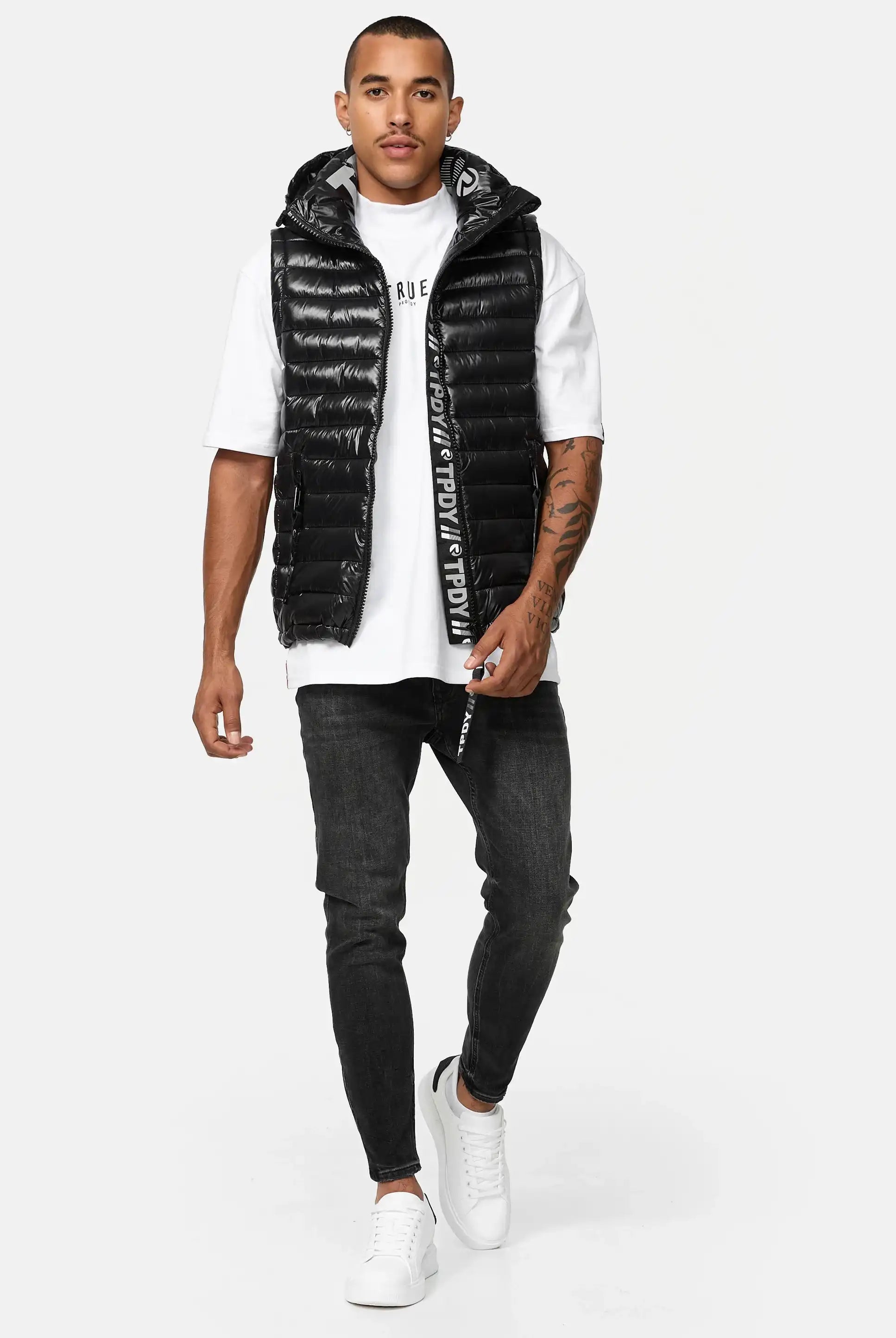 Ein Mann in einer glänzenden schwarzen Trueprodigy Jared Herrenweste mit Glanz und Kapuze über einem weißen T-Shirt, schwarzen Skinny Jeans und weißen Turnschuhen geht vor einem weißen Hintergrund nach vorne. Er hat kurze Haare, eine Tätowierung auf dem linken Arm und einen neutralen Gesichtsausdruck.