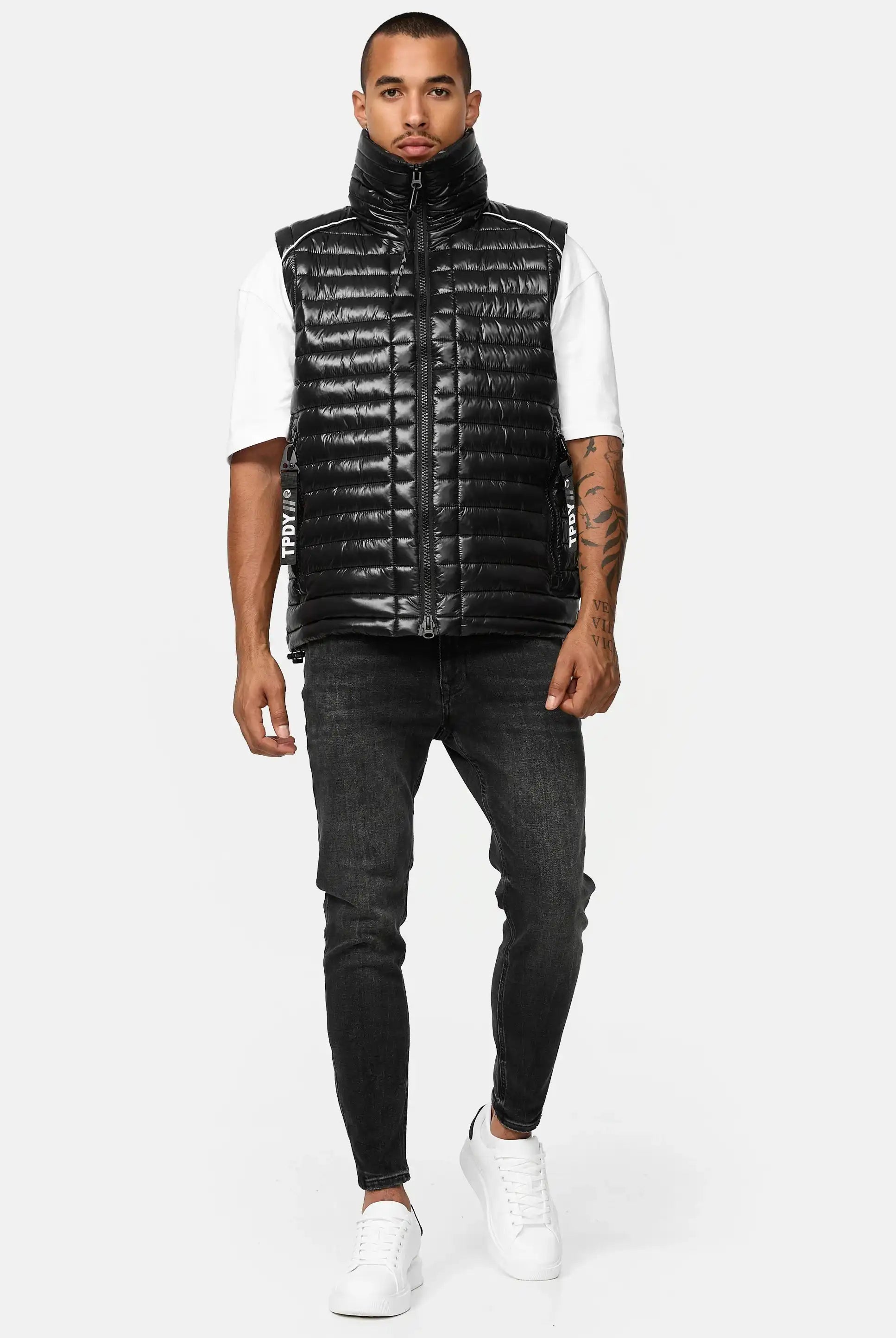 Ein Mann, der eine glänzende schwarze Trueprodigy Dave Herrenweste mit Glanz und Stehkragen über einem weißen kurzärmeligen Hemd, schwarze Skinny Jeans und weiße Turnschuhe trägt, steht vor einem schlichten hellgrauen Hintergrund.