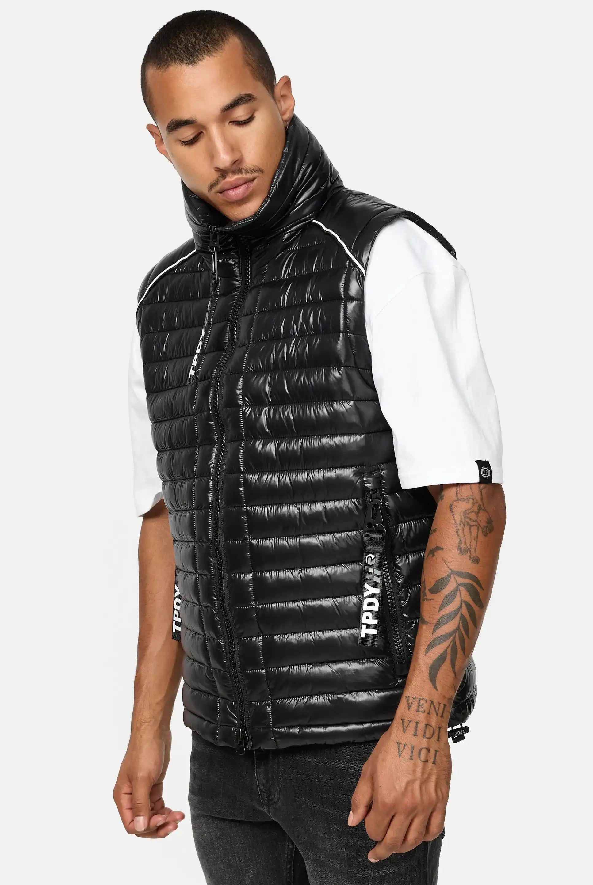 Ein Mann in einer schwarzen, glänzenden Trueprodigy Dave Herrenweste mit Glanz und Stehkragen über einem weißen kurzärmeligen Hemd steht mit leicht nach unten geneigtem Kopf. Die Weste ist mit sichtbaren Reißverschlüssen und weißem Text an den Seiten versehen.