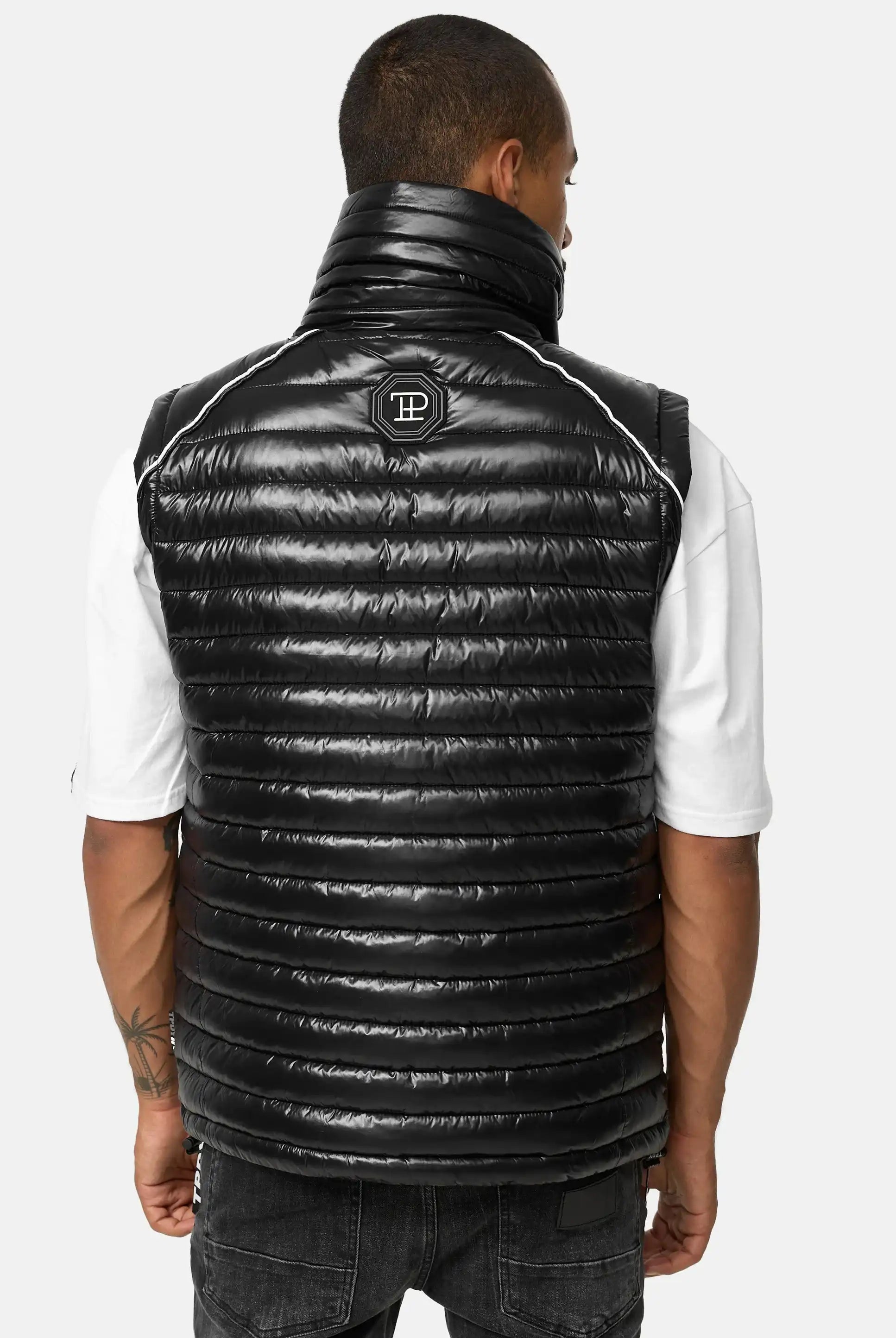 Ein Mann blickt von sich weg und trägt die trueprodigy Dave Herrenweste mit Glanz und Stehkragen mit weißem Logo auf dem oberen Rücken über einem weißen kurzärmeligen Hemd und einer schwarzen Jeans; die Weste ist horizontal gesteppt und hat einen hohen Kragen.