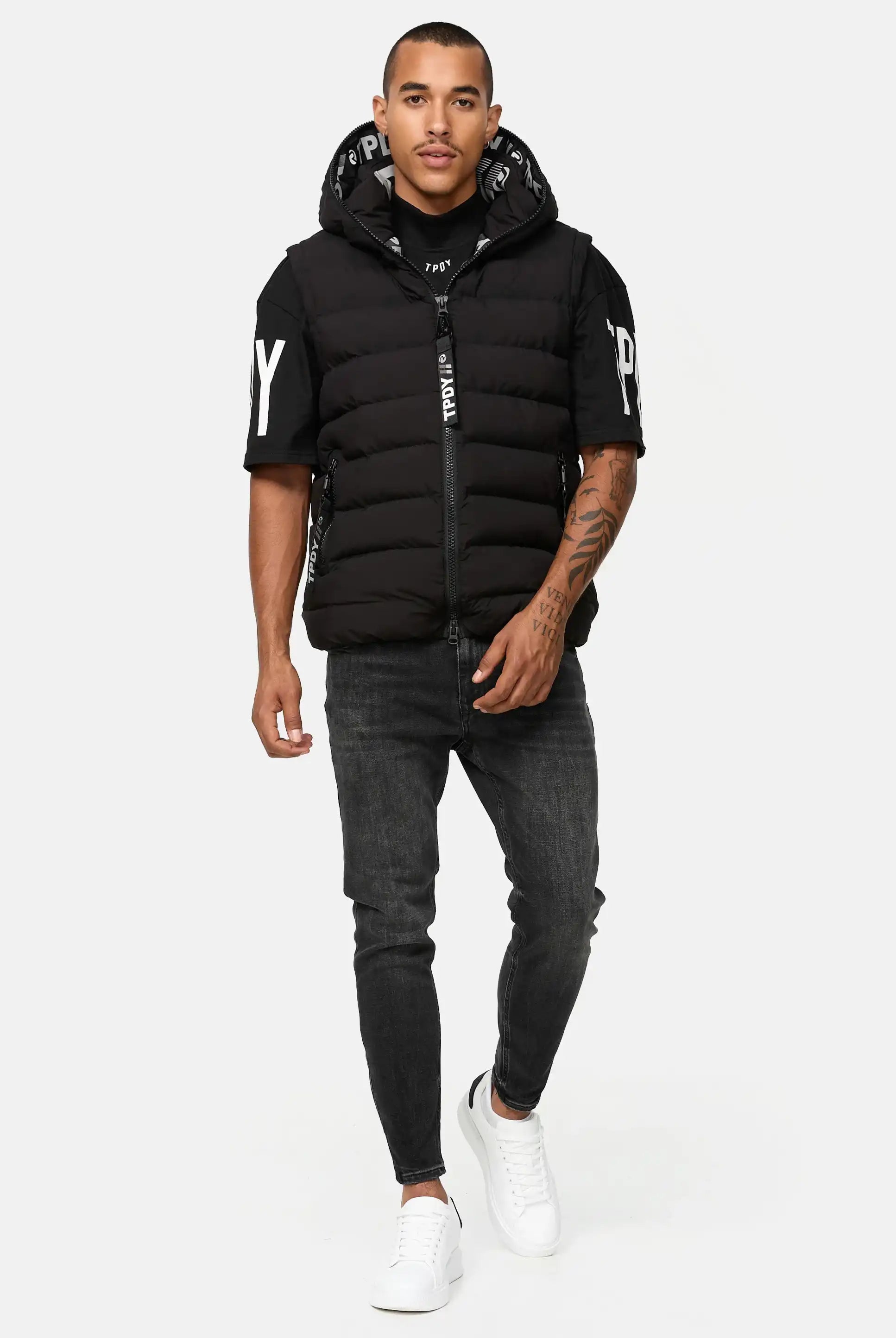 Ein Mann steht und geht nach vorne und trägt eine Trueprodigy Matthew Pufferweste mit Kapuze matt über einem schwarzen T-Shirt mit weißer Aufschrift, dunkelgraue Jeans und weiße Turnschuhe vor einem schlichten hellen Hintergrund.