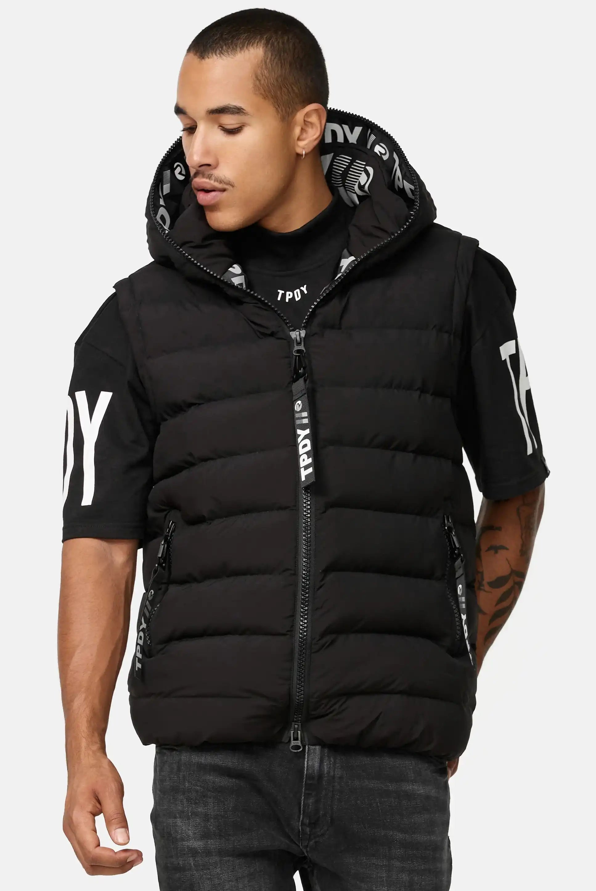 Ein Mann trägt die Trueprodigy "Matthew Pufferweste mit Kapuze matt" - eine schwarze Kapuzenweste mit Reißverschluss und aufgedruckten Textdetails - über einem schwarzen T-Shirt mit weißer Aufschrift und steht mit leicht zur Seite gedrehtem Kopf.